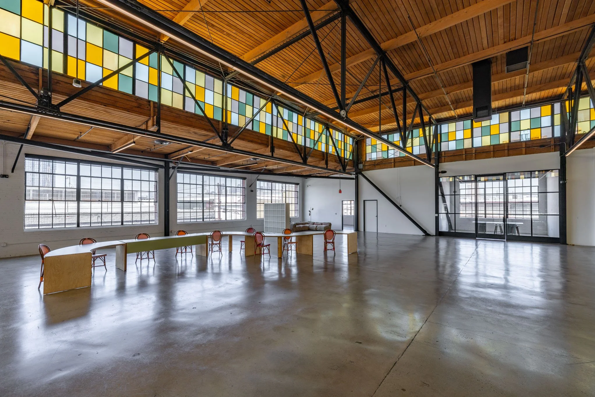 Spacious industrial-style event space with large windows, colorful stained glass upper windows, wooden ceiling, and minimal furniture including a curved table with red chairs and a small sofa.