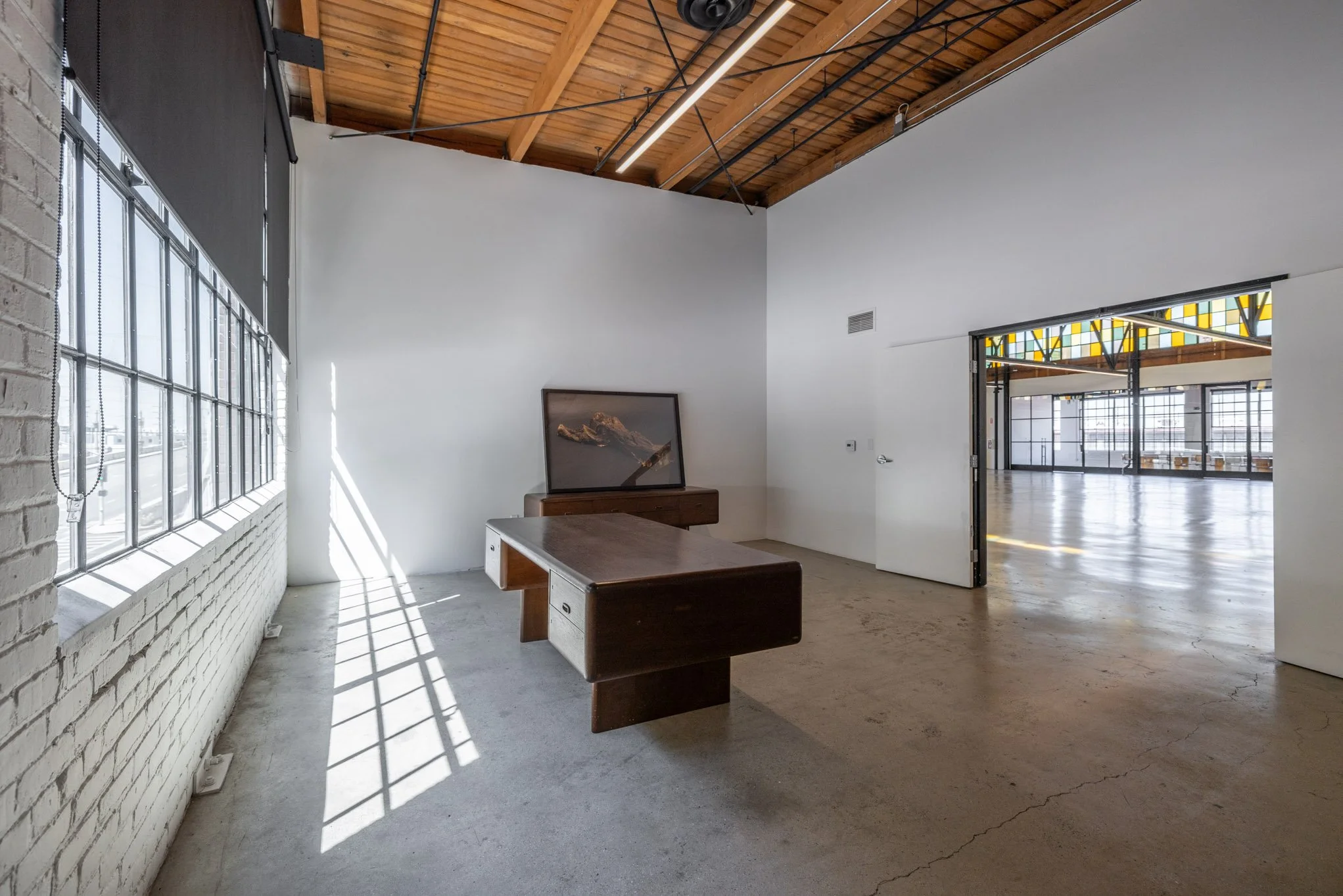 Interior of a spacious, industrial-style room with white walls, a wooden ceiling, large windows, a wooden desk, and a picture leaning against the wall