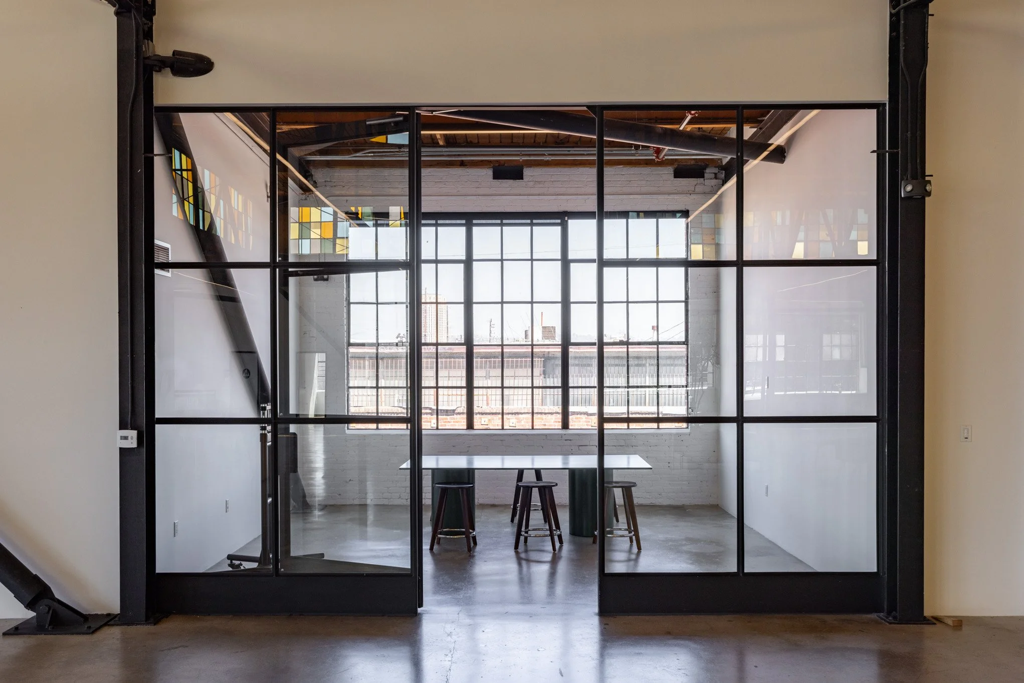 Modern office meeting room with glass door and large window, three chairs, and a tall table, with natural light and exposed ceiling beams.