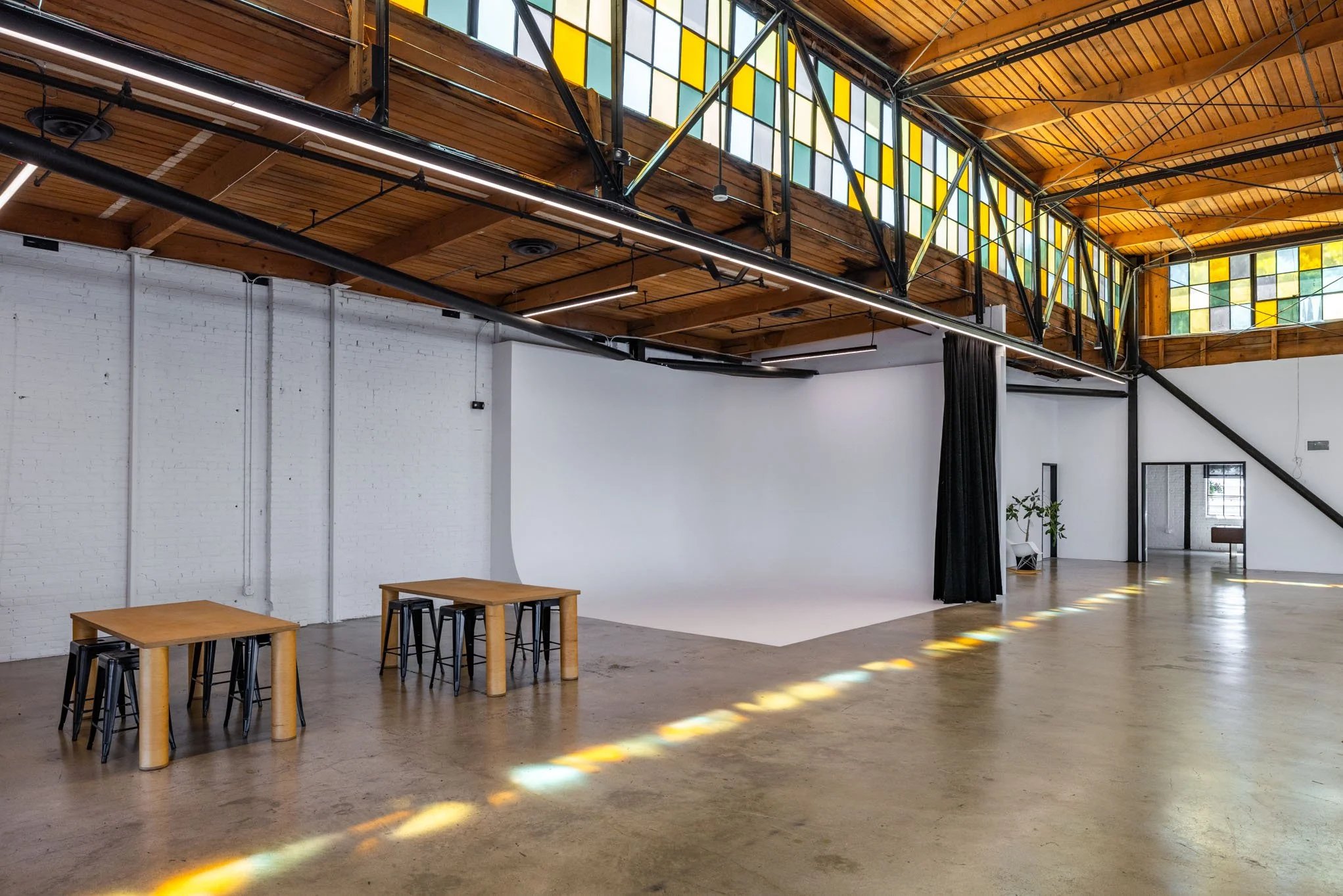 An empty photography studio with a white backdrop, wooden tables and black stools, a staircase, stained glass windows, and polished concrete floors.