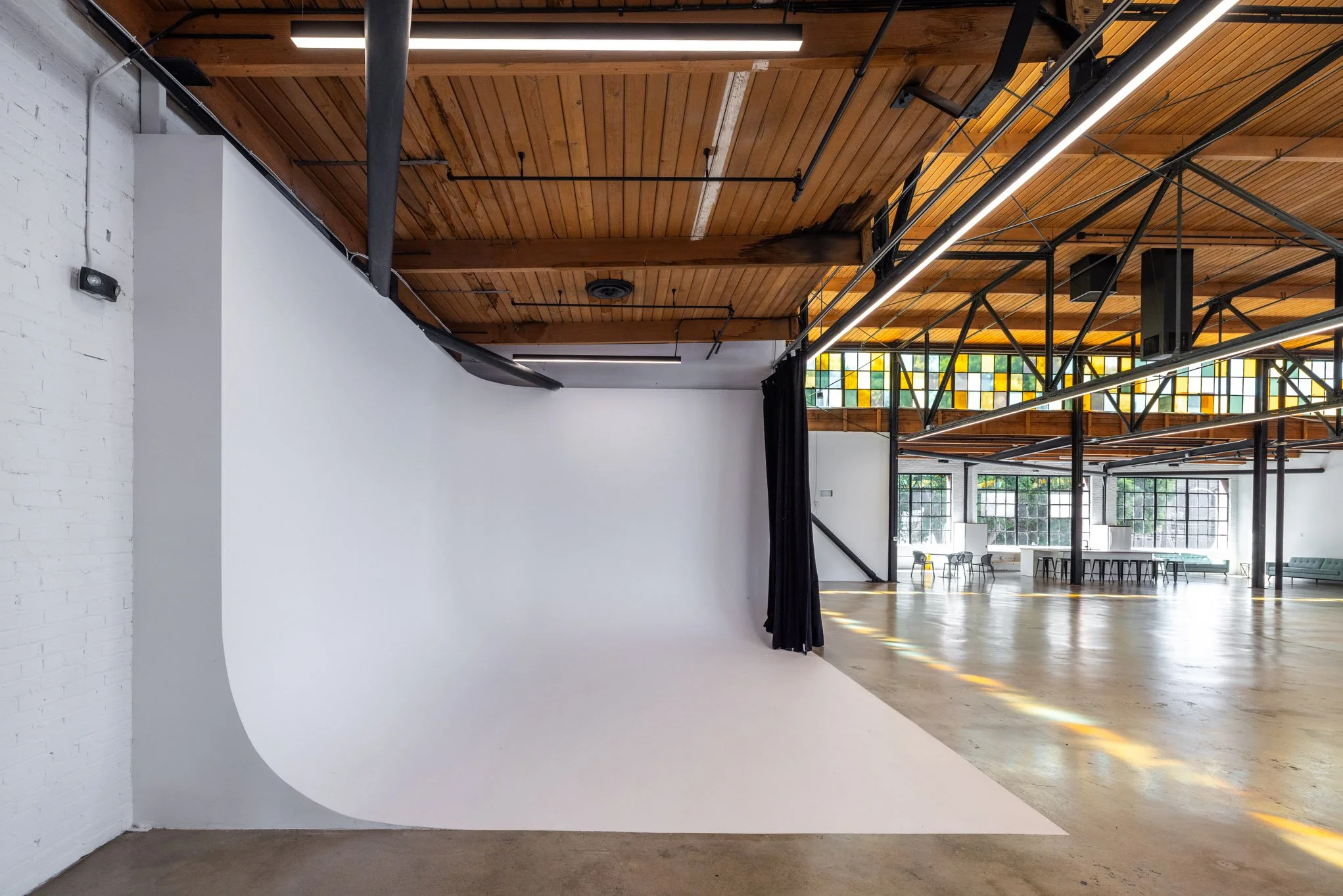 Empty photography studio with a white seamless backdrop, black curtains, and large windows in a spacious room with wooden ceiling and exposed rafters.