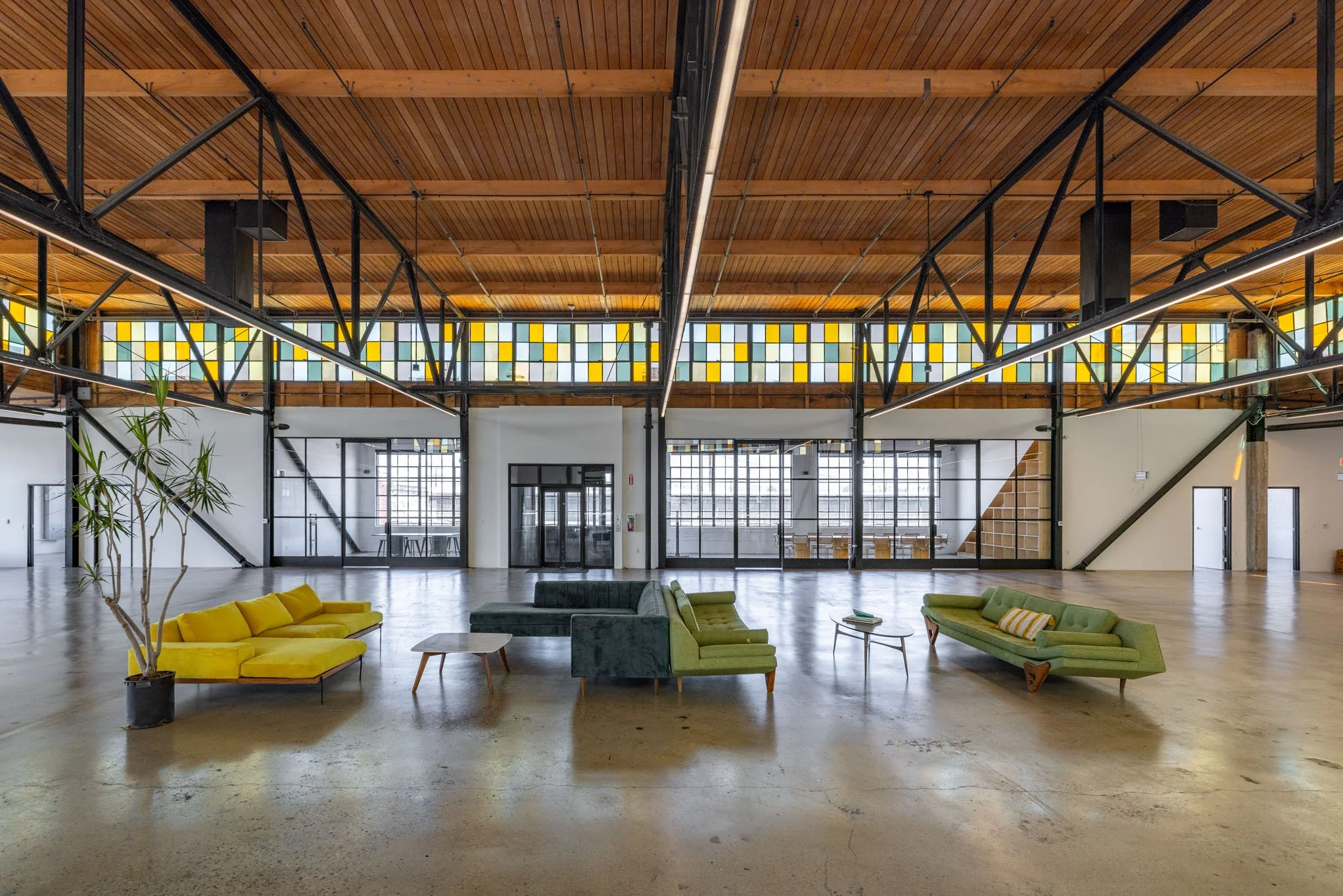 Open spacious lounge area with colorful sofas, small tables, potted plant, and high ceiling with wooden beams and multicolored window panels.