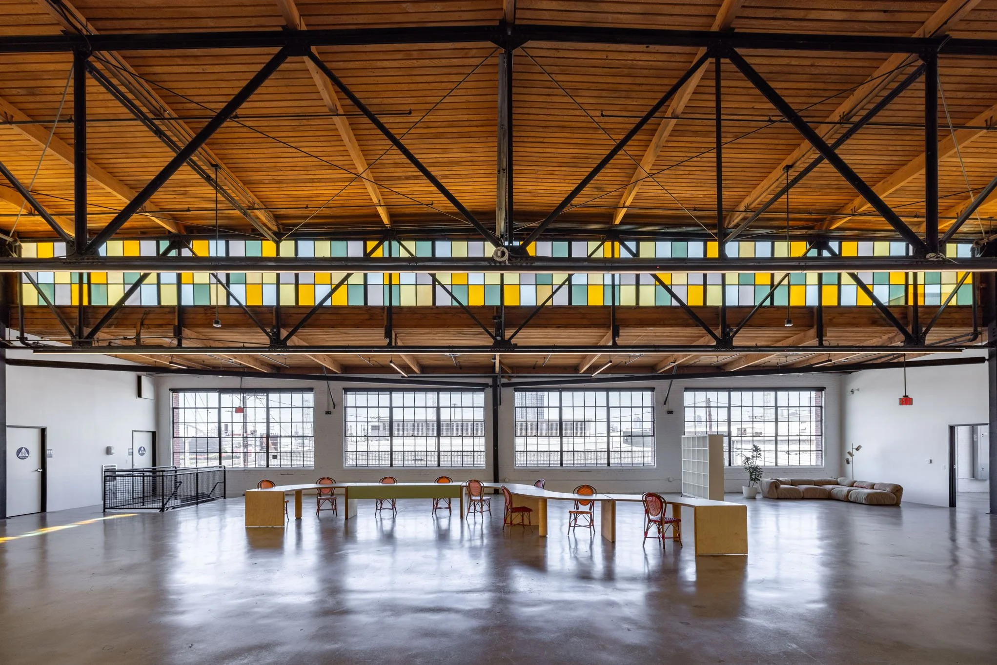 Interior of a modern, spacious room with large windows, wooden ceiling with black metal beams, colorful stained glass window strip, minimal furniture including chairs, a sofa, and a partition.