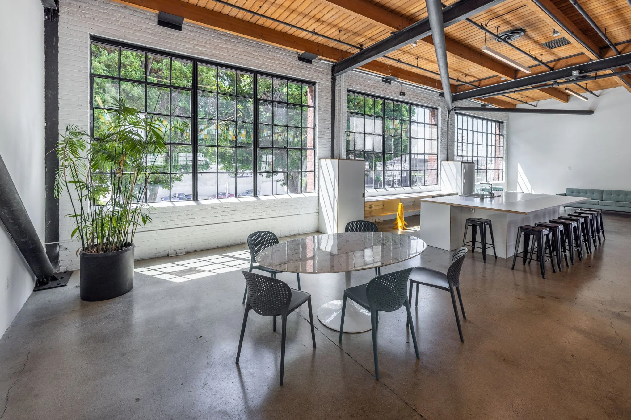Modern industrial-style interior with large grid windows, concrete floors, a round marble table with four chairs, a kitchen island with black stools, a large potted plant, and a corner seating area with a mint green couch.