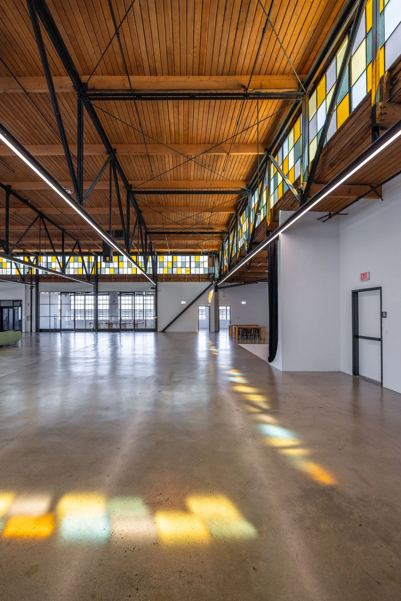 Empty, modern open space with concrete floors, wooden ceiling, large stained glass windows, and minimal furnishings.