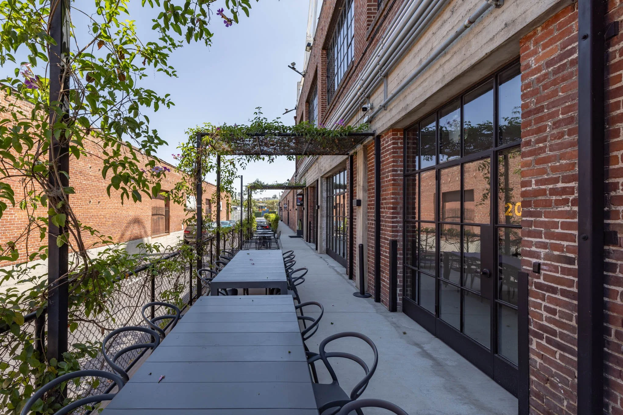 An outdoor seating area with black tables and chairs under a trellis on a sidewalk outside a brick building.
