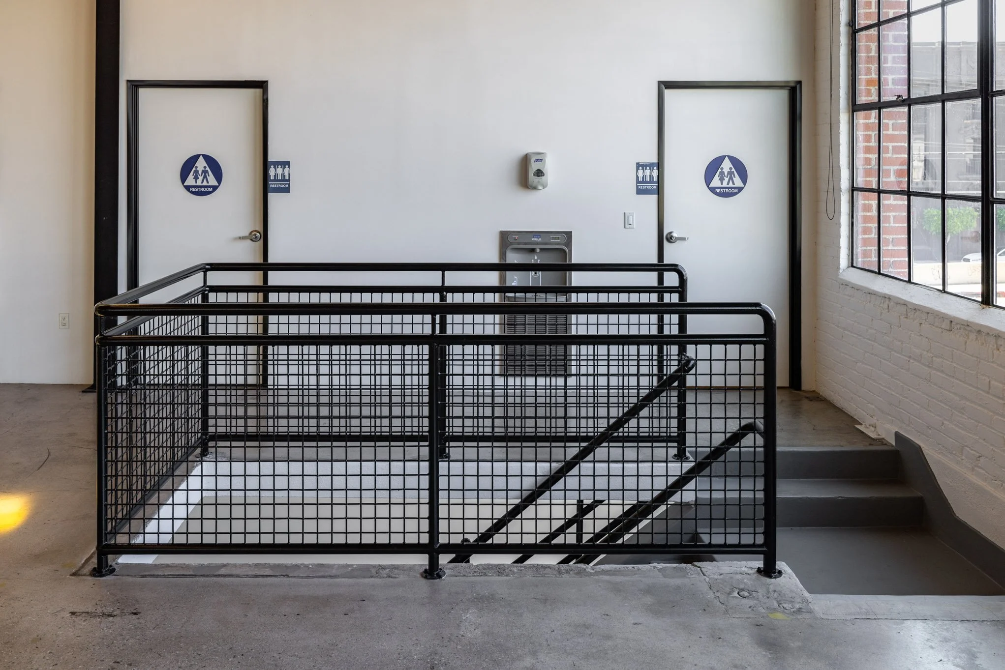 Indoor staircase with black metal railing, leading down to a basement or lower level, with men and women restrooms doors on the wall at the top of the stairs and an automatic hand sanitizer dispenser in between, next to a water fountain, near a large