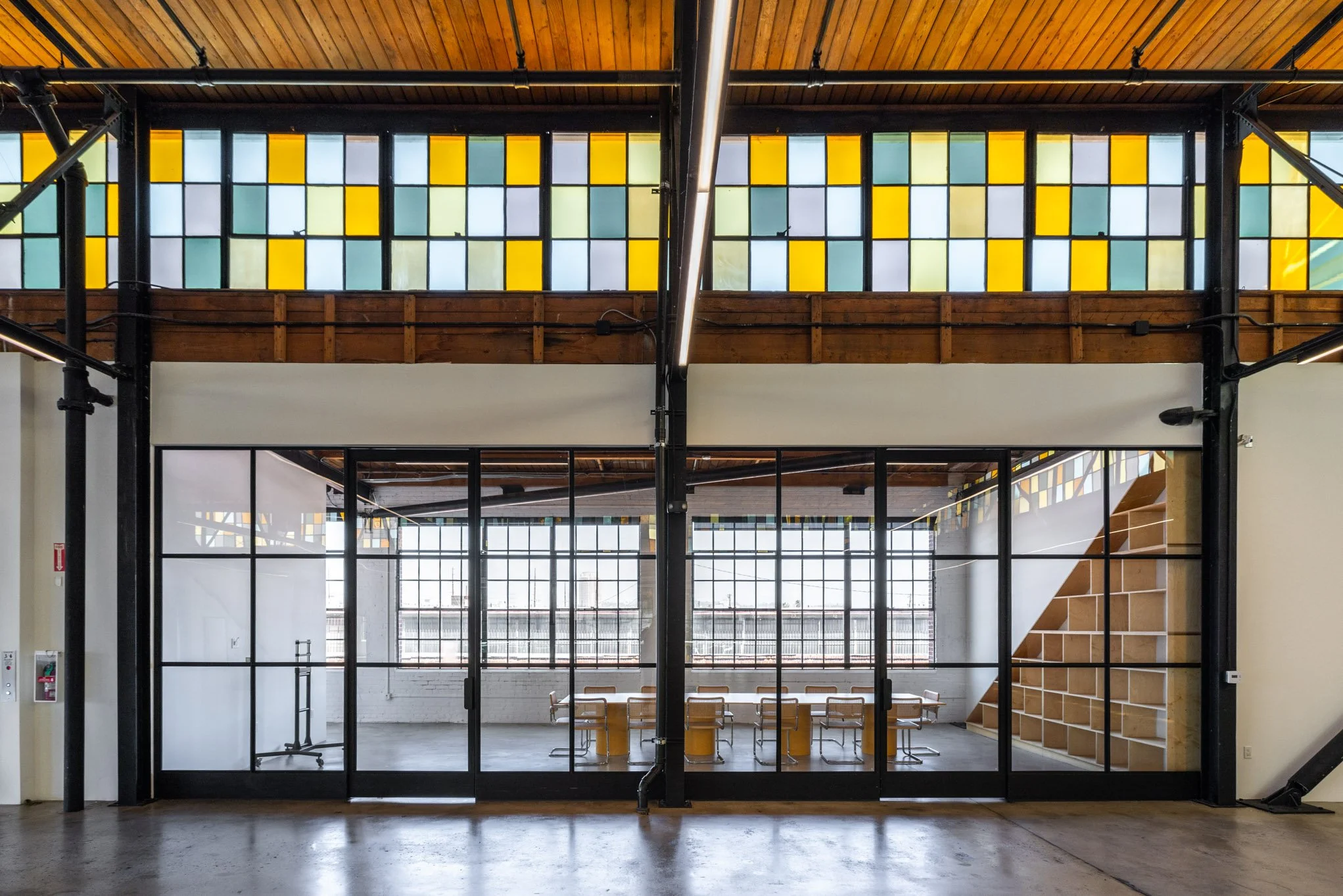 Interior view of a modern office space with glass walls, a wooden staircase, and large stained glass windows with a yellow, blue, and green pattern, high ceiling with wooden panels.