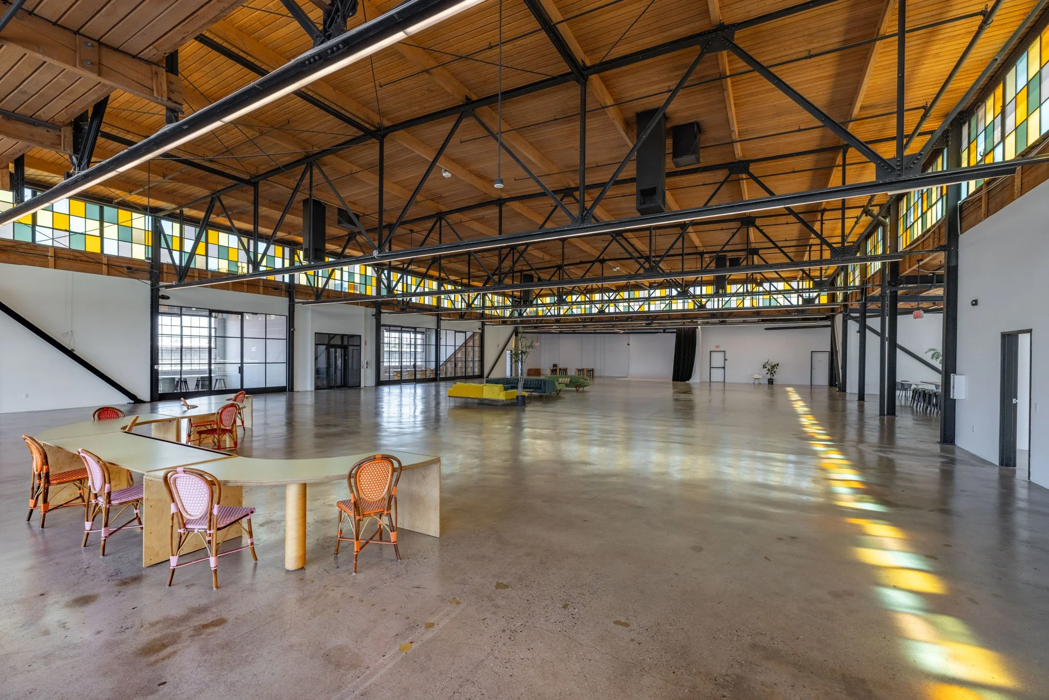 Large empty event space with high wooden ceiling, black metal beams, stained glass windows, and scattered furniture including chairs and couches.