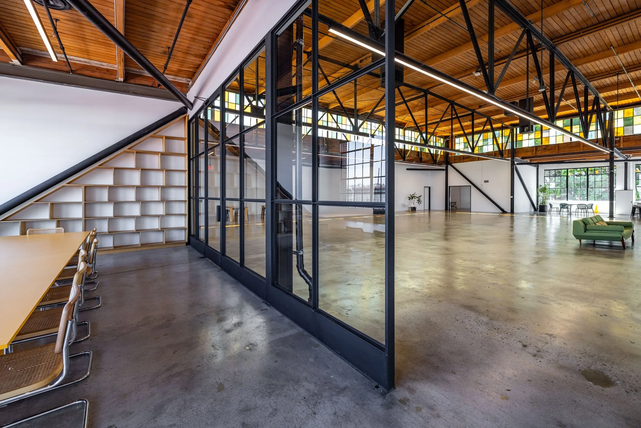 Open, modern industrial-style office space with glass partition walls, wooden ceiling beams, and minimal furniture including a green sofa, chairs, and tables.