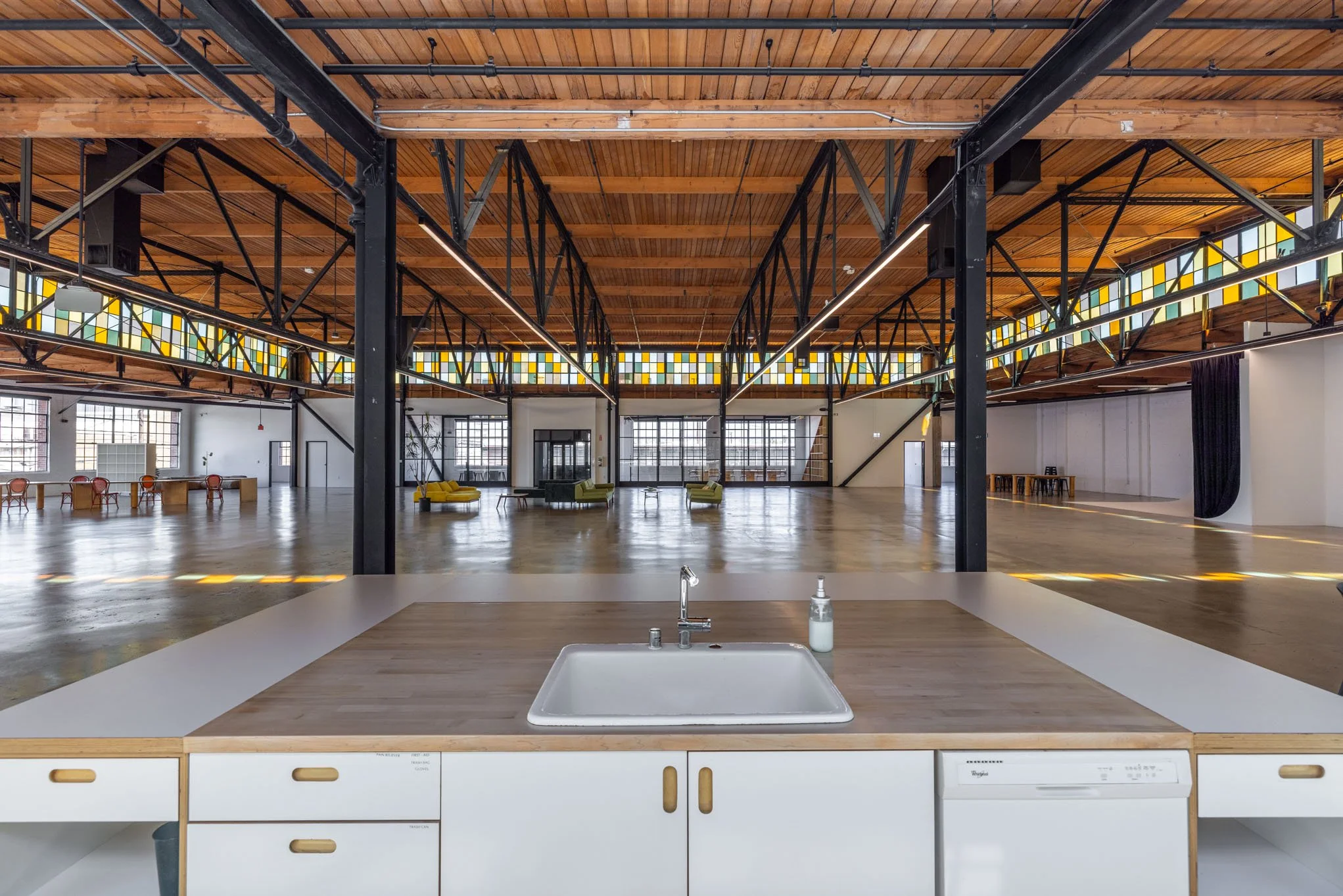 Open, spacious industrial-style loft with high wooden ceilings, exposed black metal beams, large windows, and colorful seating areas, viewed from the kitchen counter with sink and soap dispenser.