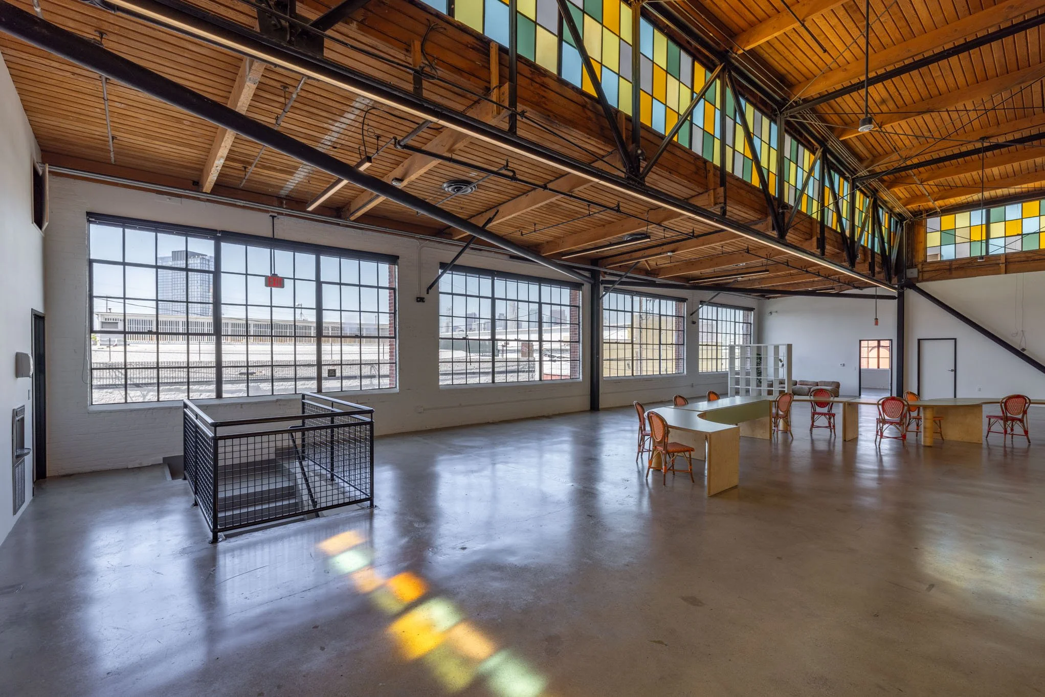 Spacious industrial-style room with large windows, high wooden ceiling, colorful stained glass windows near the ceiling, and minimal furniture including a table, chairs, and a shelving unit.