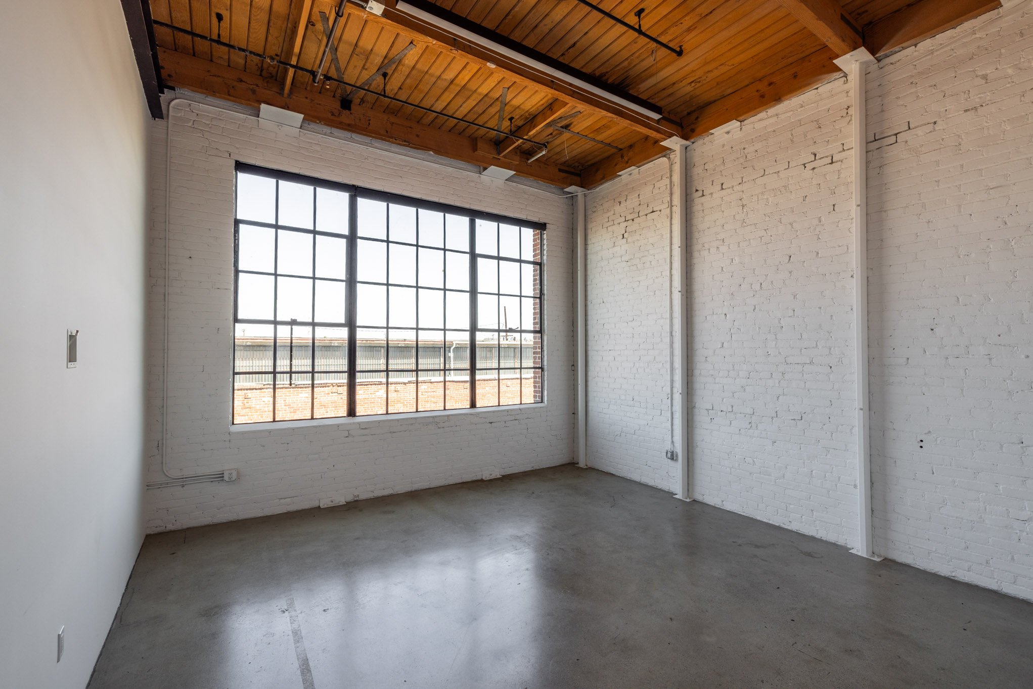 Empty room with white brick walls, large window with grid panes, concrete floor, and wooden ceiling.