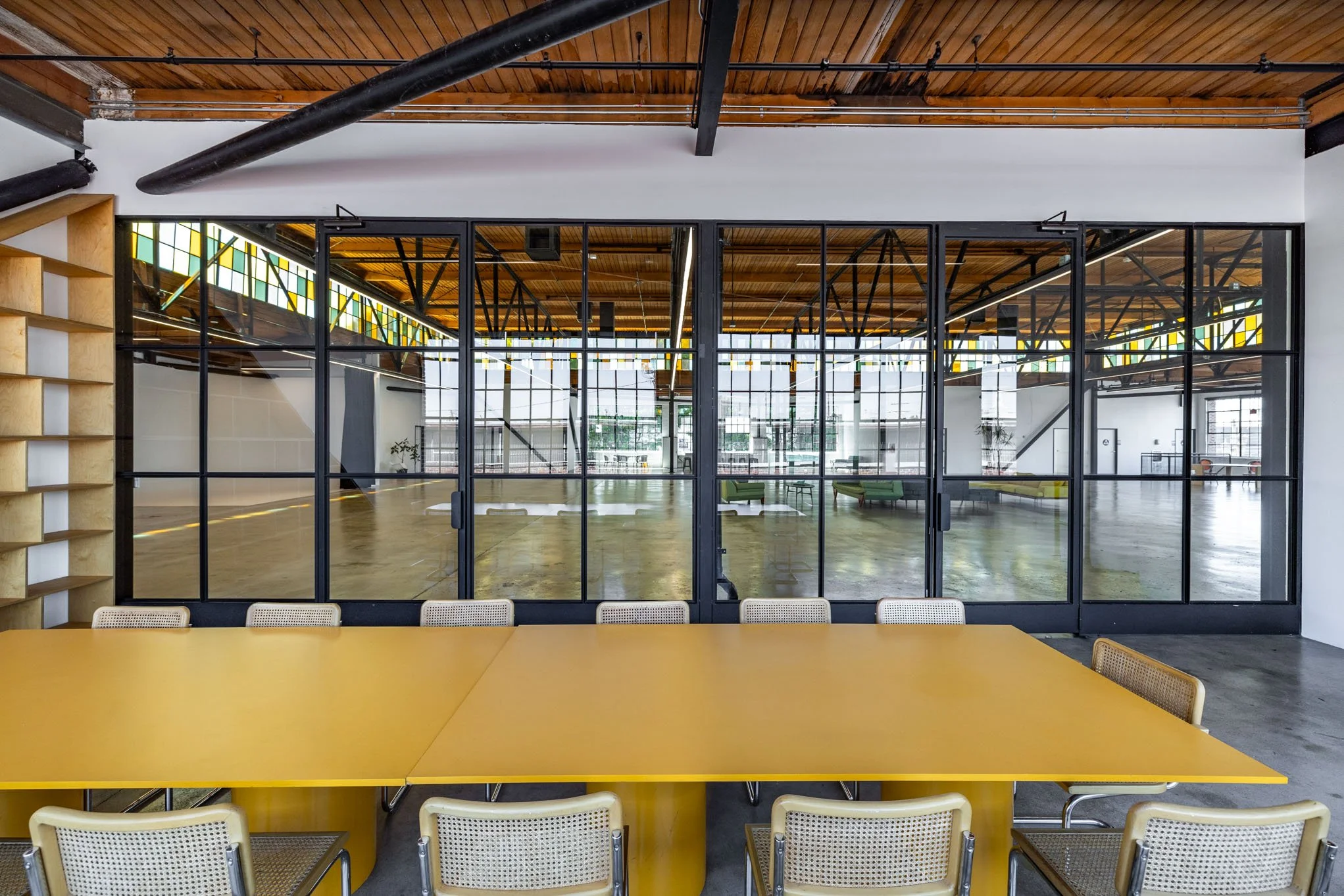 Conference room with a long yellow table and beige chairs, large glass sliding doors, open office with exposed wooden ceiling and industrial black pipes, stained glass windows.
