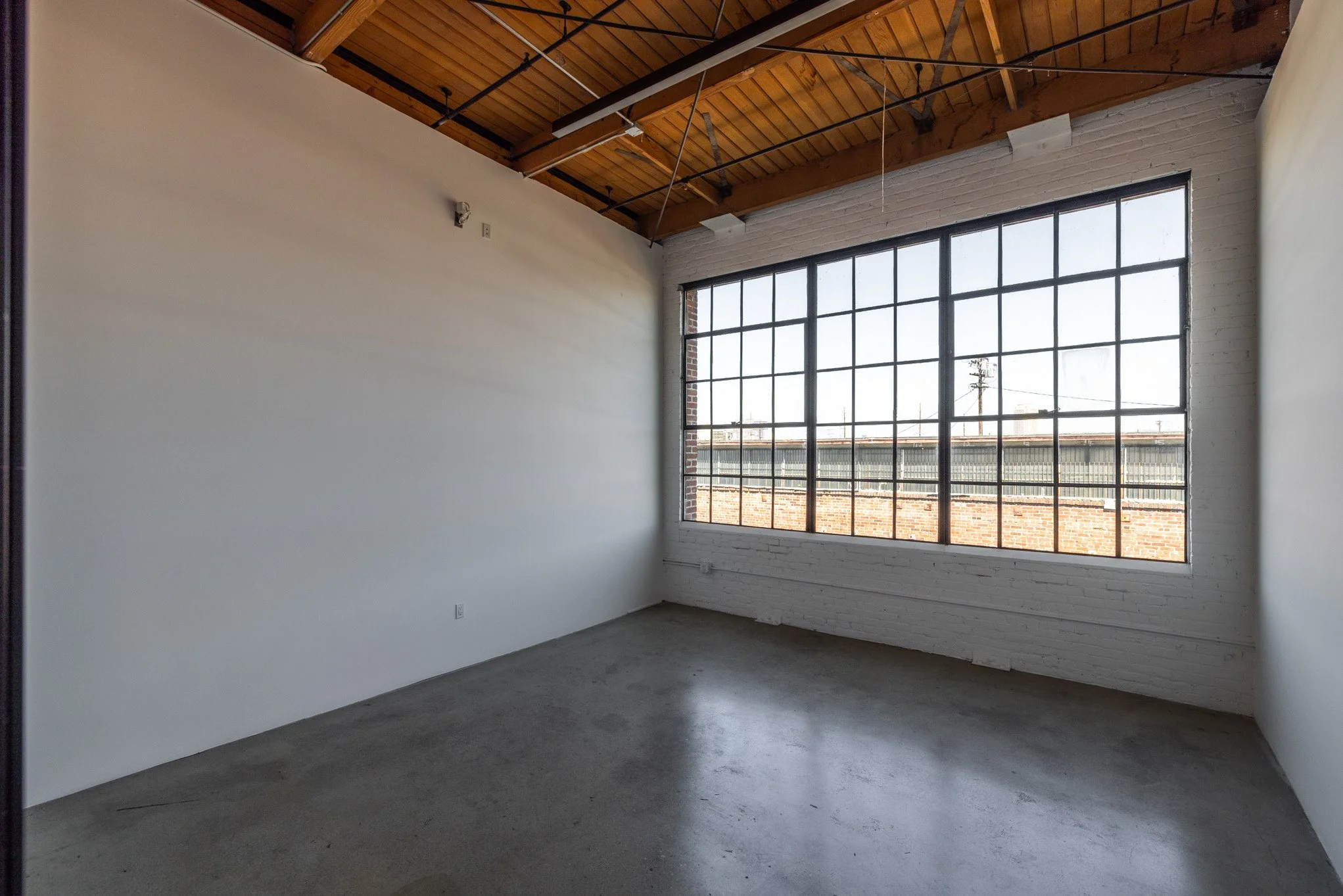 Empty room with concrete floor, white brick wall, large window with grid pattern, wooden ceiling with exposed beams, and a view of rooftops and sky outside.