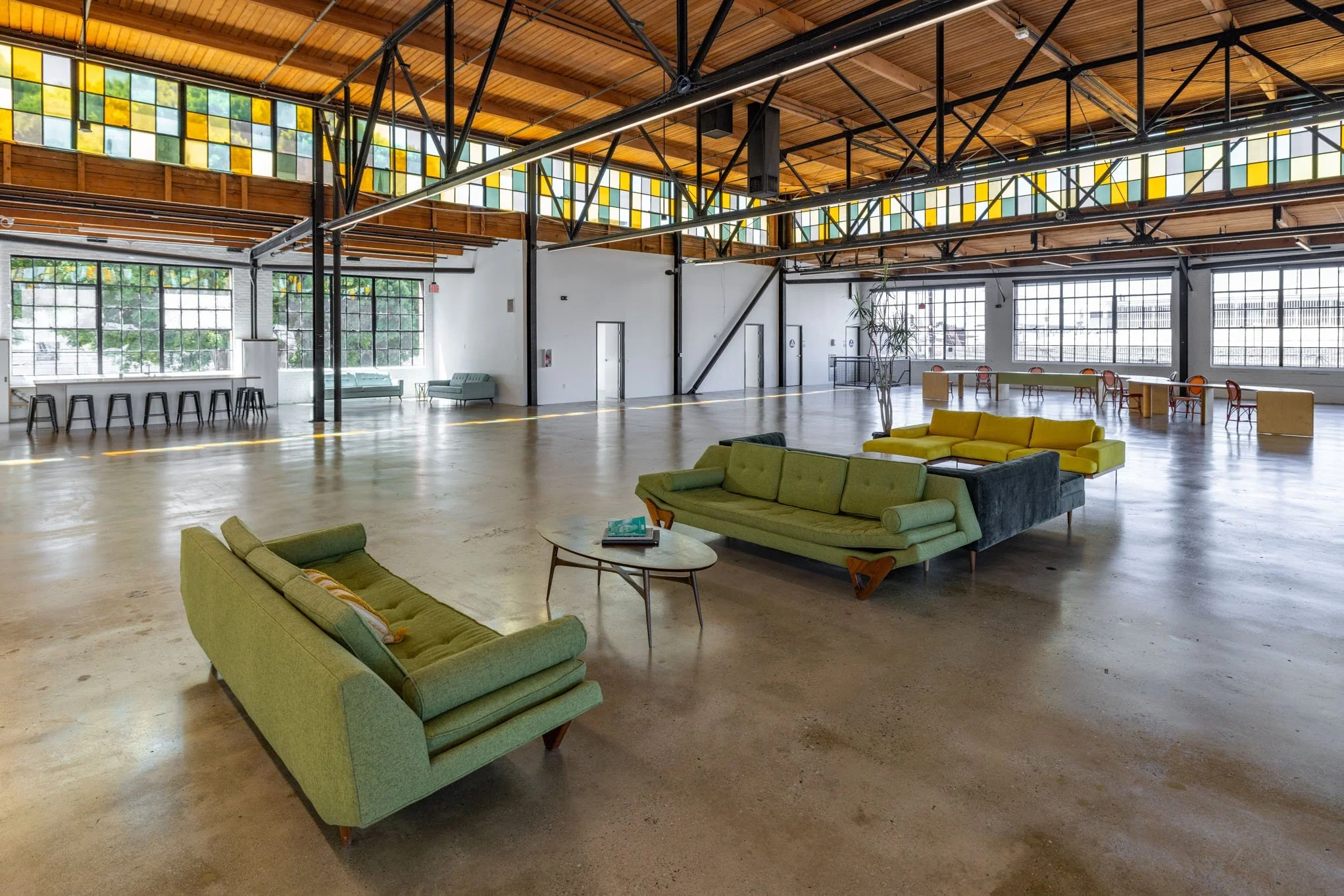 Spacious, open-plan industrial-style interior with vintage green and yellow sofas, a round wooden coffee table, and high windows with colorful stained glass accents.