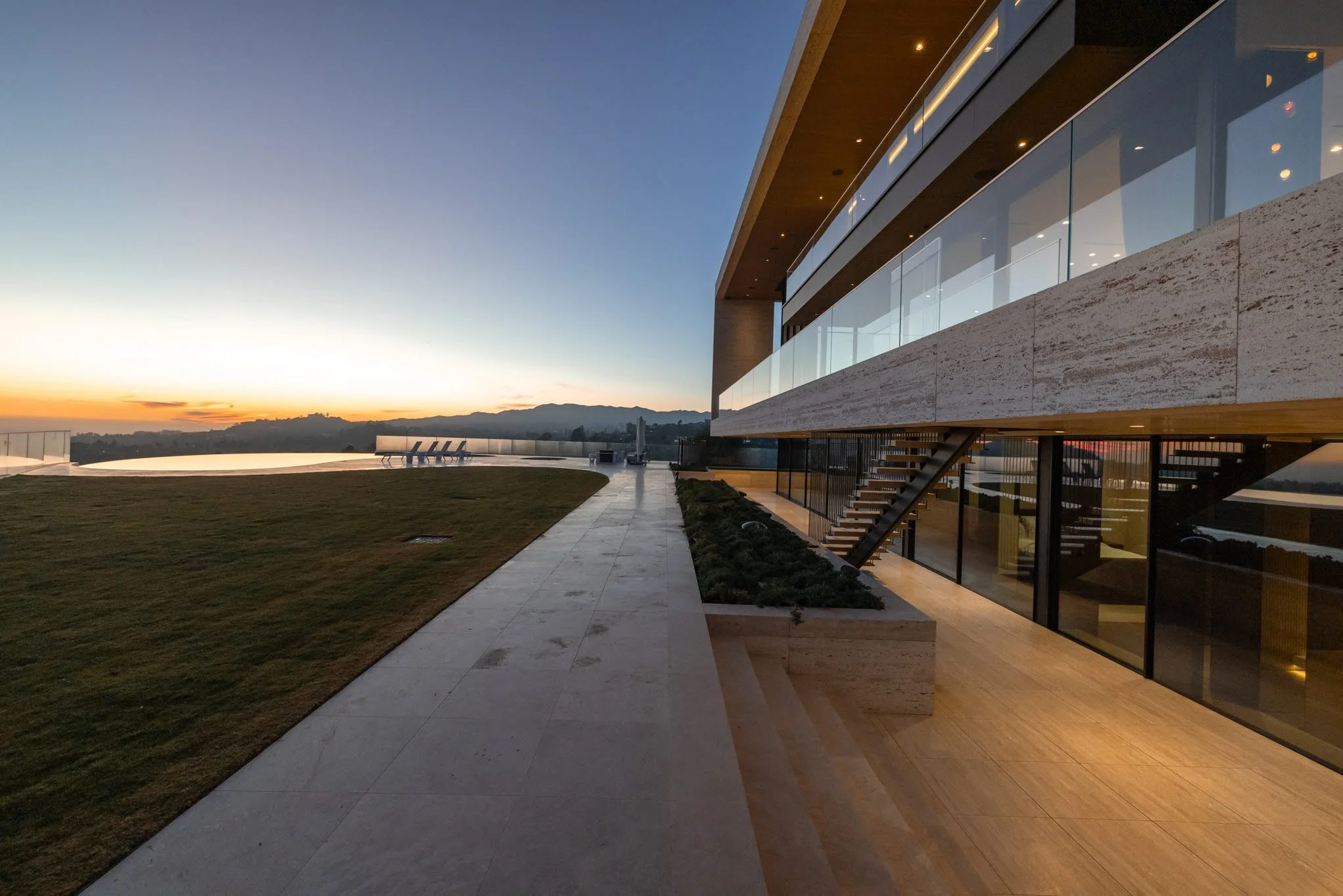 Modern building with glass balconies and stairs, overlooking a grassy area and an infinity pool at sunset.