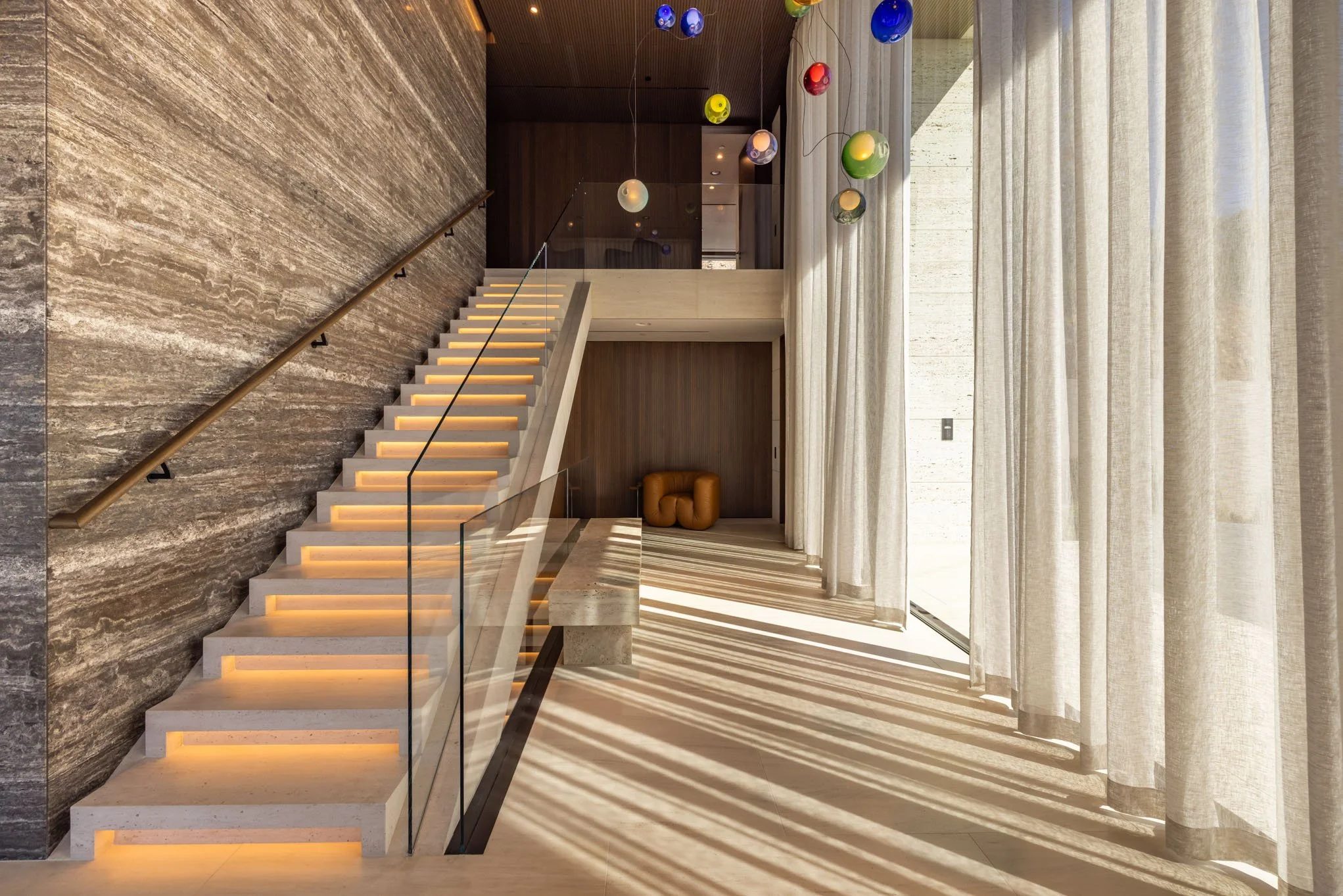Modern staircase with illuminated steps, glass railing, and a wall with textured stone finish. Large window with floor-to-ceiling curtains casting shadows on the floor, and colorful hanging pendant lights.