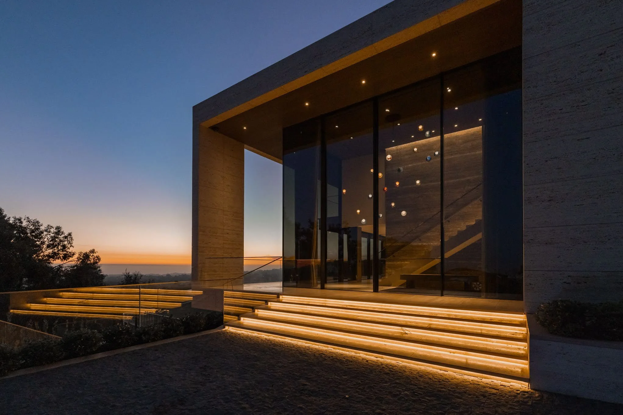 Modern house with large glass windows and illuminated stairs at sunset.