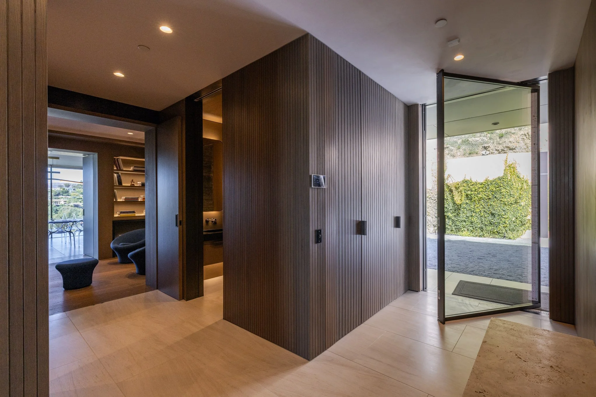 Modern interior entrance with wooden panel walls, a glass door leading outside, and a sitting area with chairs and bookshelves in the background.