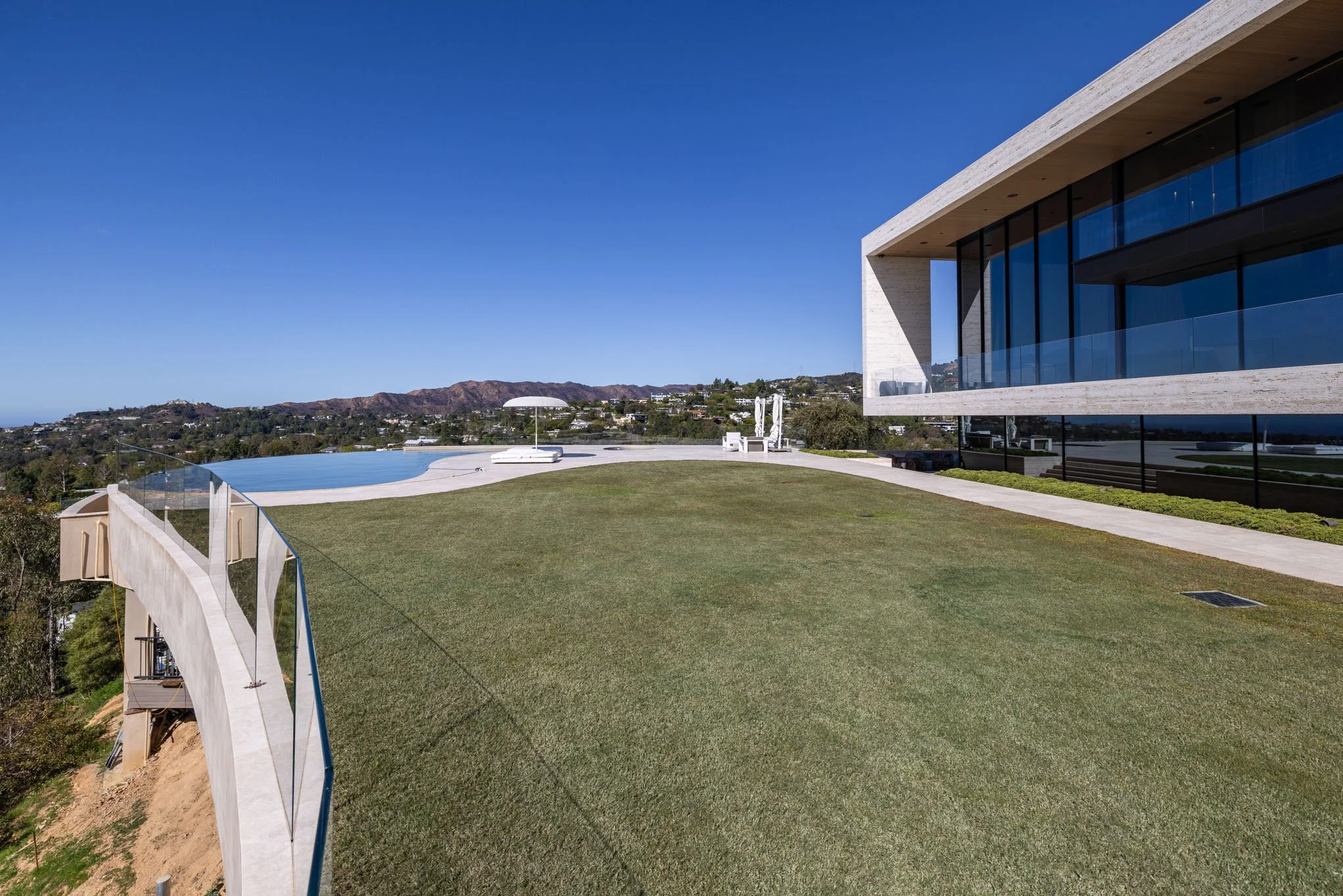 Modern house with large glass windows, a swimming pool, metal and glass railing, outdoor seating, and a well-kept lawn on a clear sunny day.