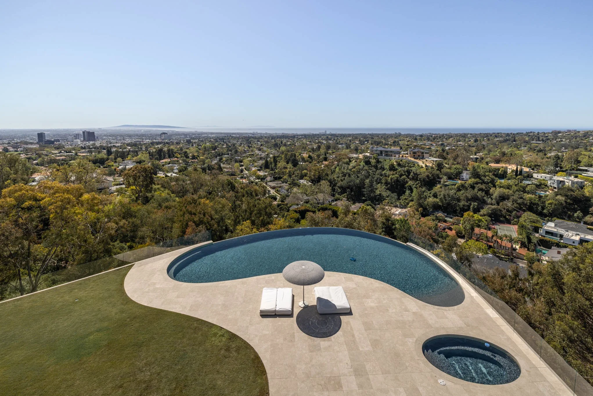 A rooftop pool with lounge chairs and an umbrella overlooking a cityscape with trees, buildings, and distant mountains on a clear day.
