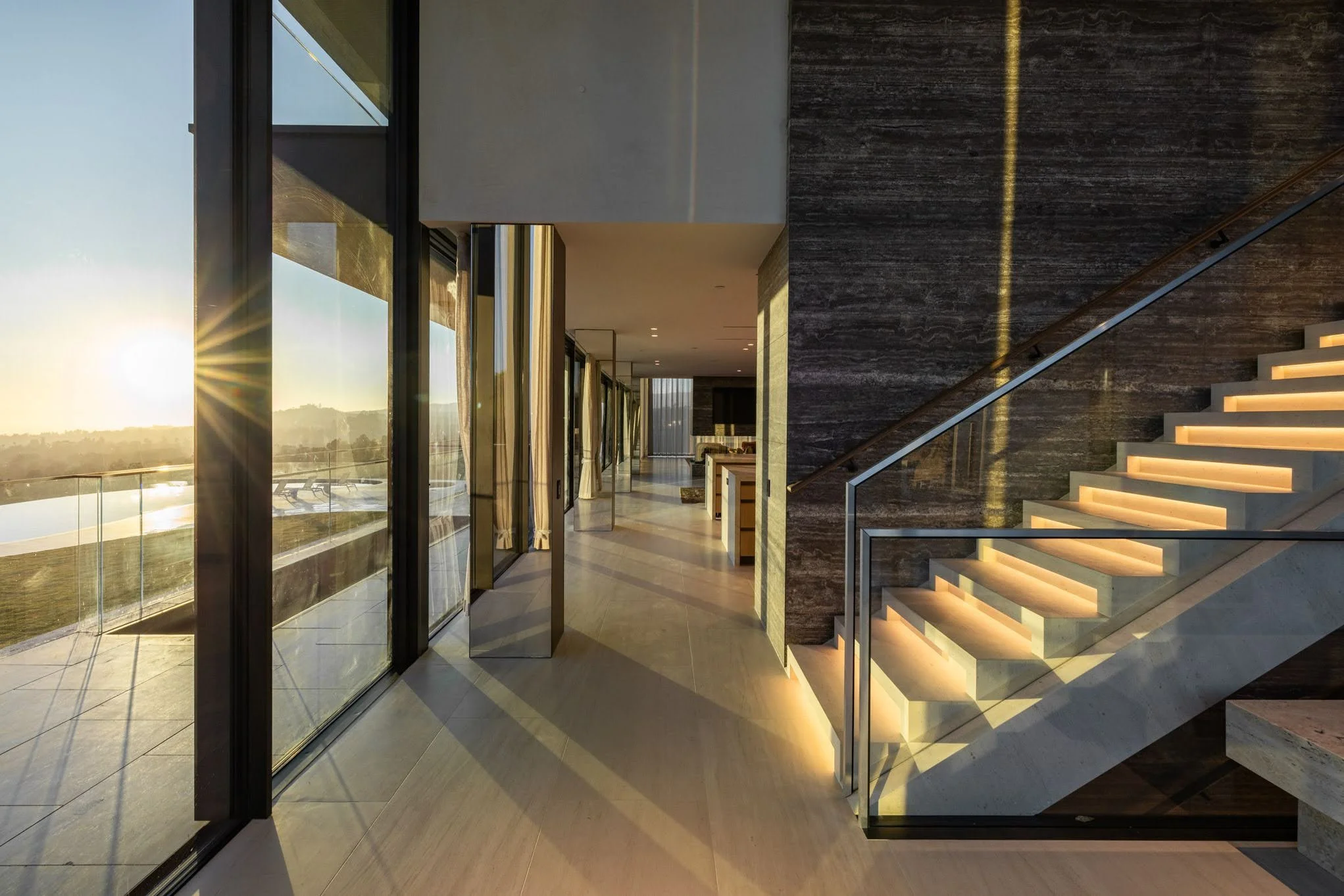 Sunset view from the balcony of a modern house with glass doors, wooden floor, and a staircase with lit steps.