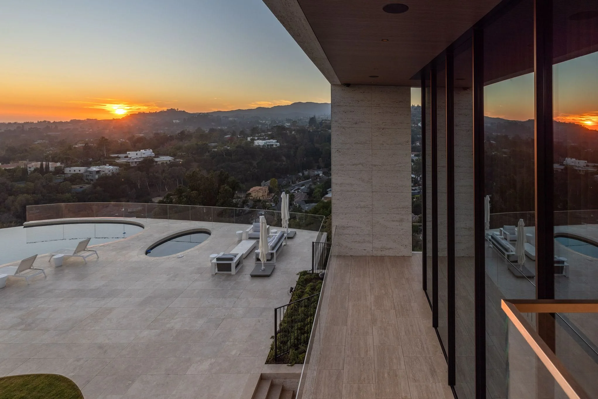 A modern balcony with a view of a sunset over hills, featuring a swimming pool, lounge chairs, umbrellas, and glass walls reflecting the sunset.