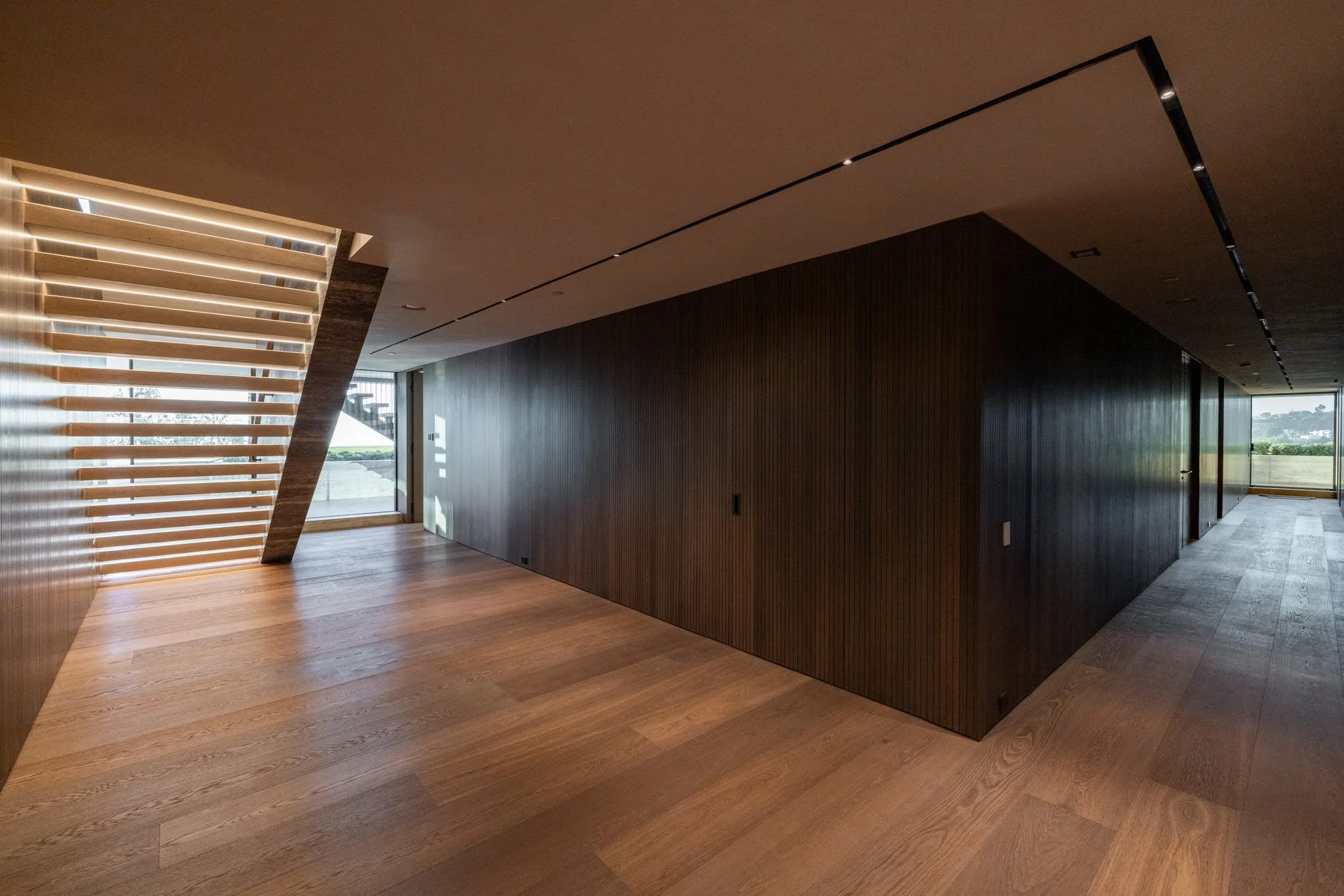 Interior of a modern home with wooden floors, dark wood-paneled walls, and a staircase with horizontal slats near a large window.