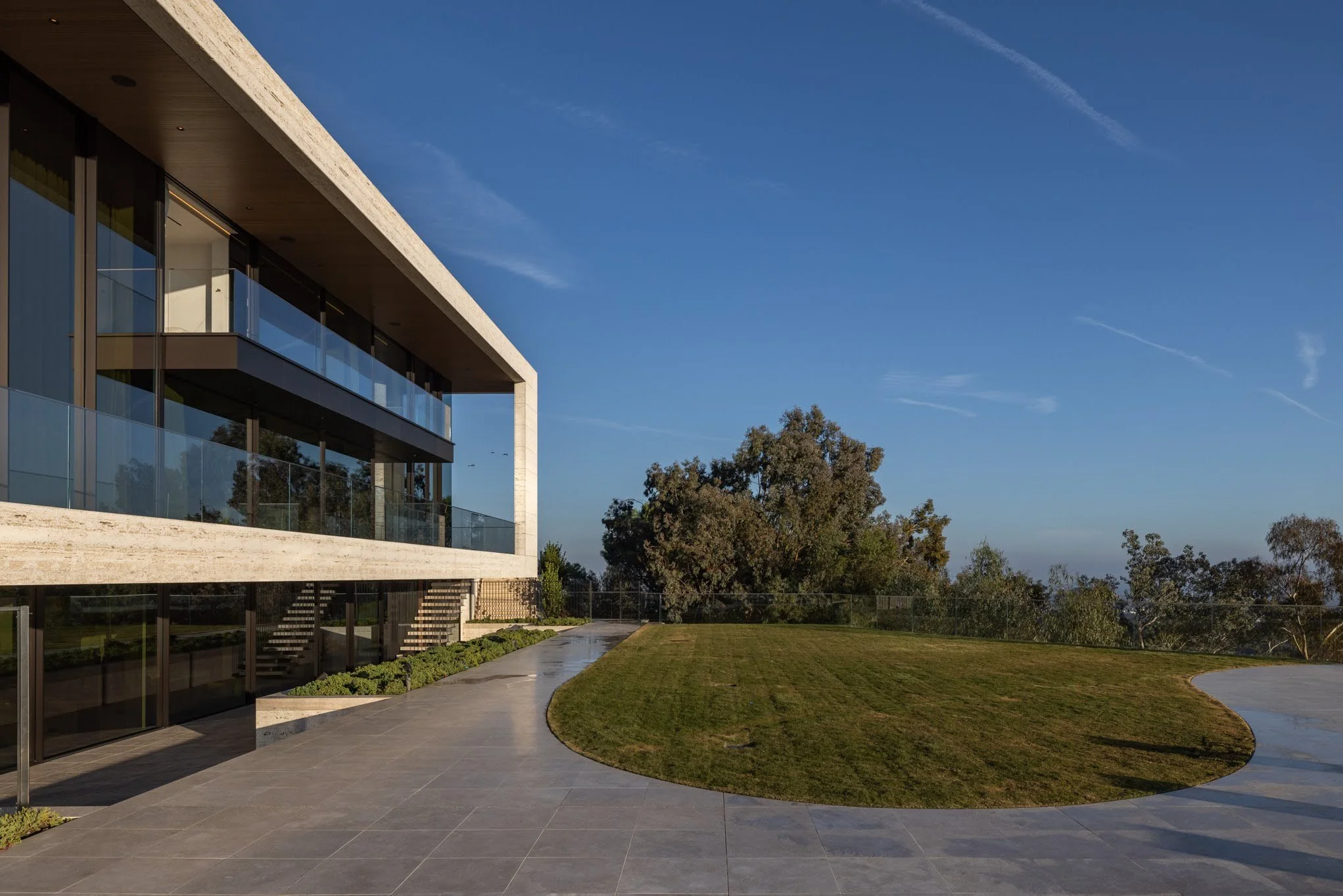 Modern residential building with large glass windows and balconies, a paved patio area, and a grassy yard, with trees and a blue sky in the background.