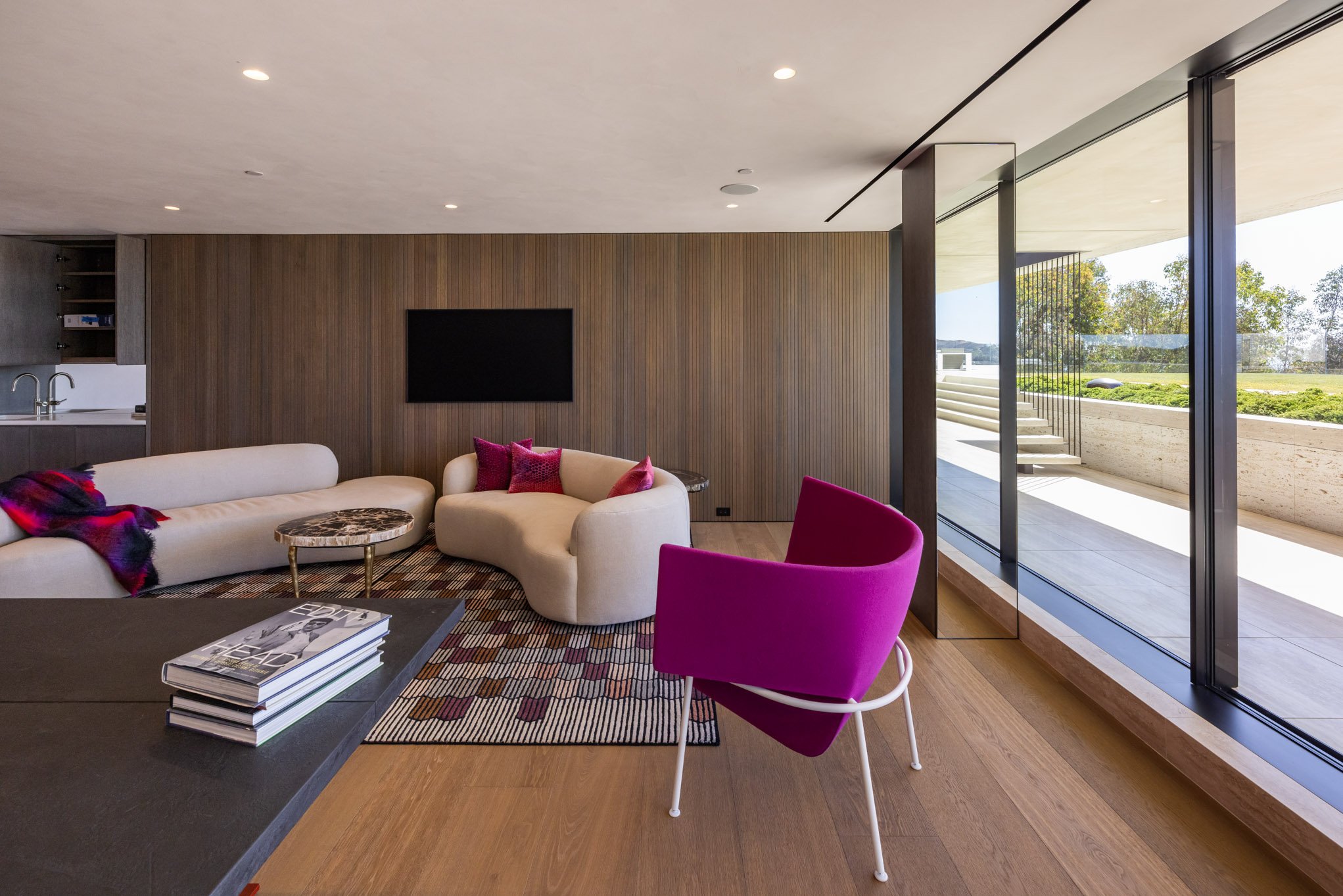 Modern living room with beige and magenta furniture, wooden wall, flat-screen TV, books on table, large glass sliding doors with view of outdoor patio and greenery.