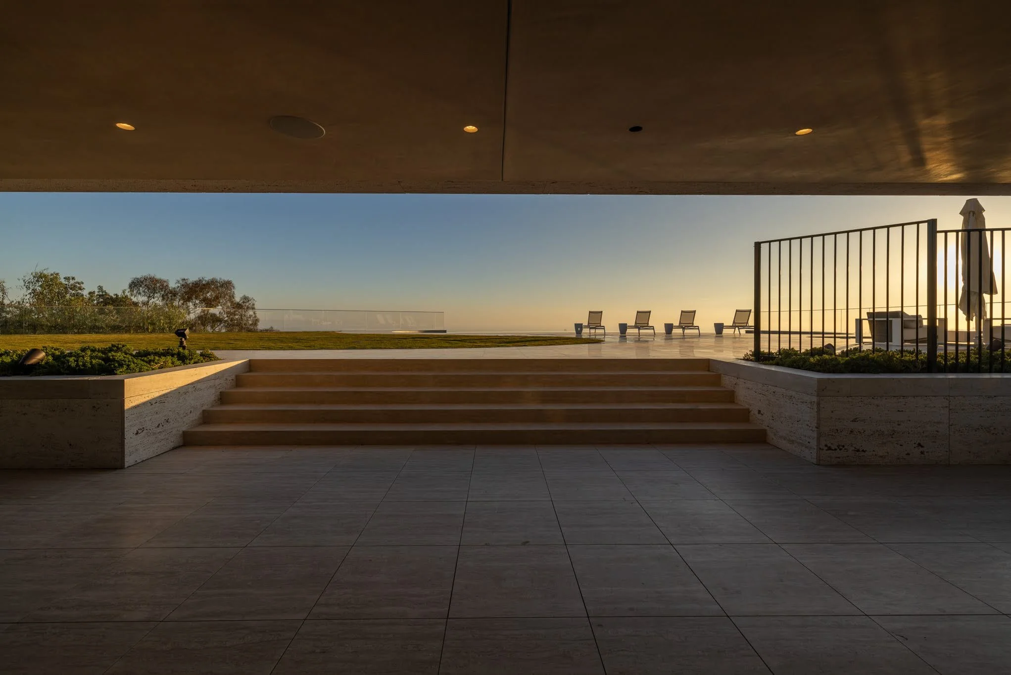 View of a modern outdoor patio with steps leading up to a grassy area and lounge chairs facing a sunset horizon, framed by a concrete overhang and a metal fence.