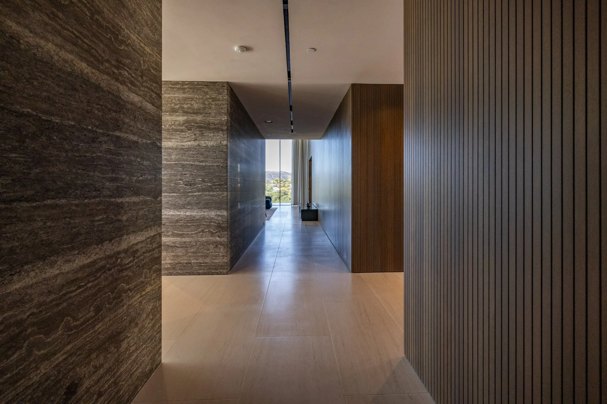 Modern hallway with stone, wood, and dark panel walls leading to a spacious room with large window and scenic view.