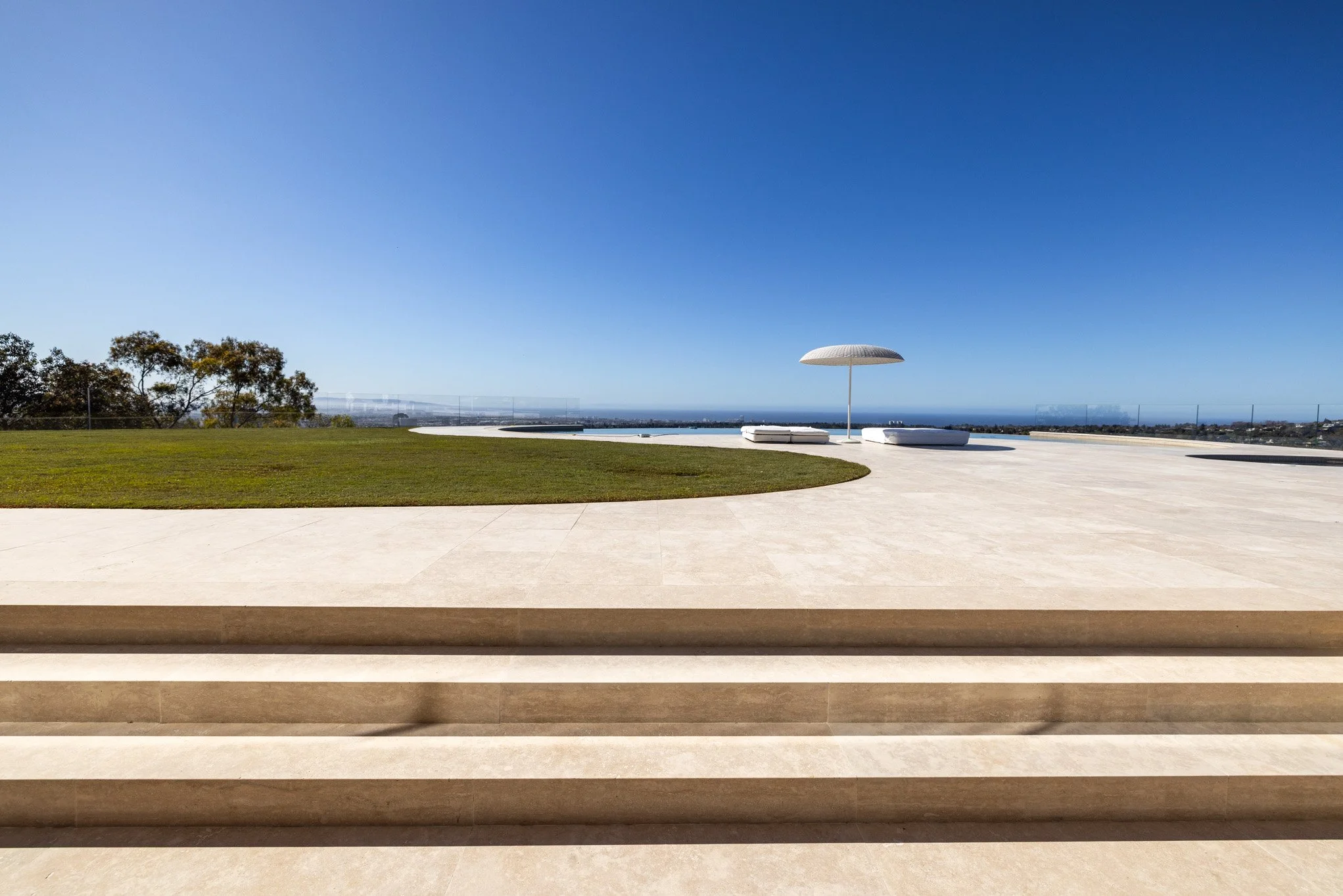 View of an outdoor patio with steps leading down to a grassy area, a large white umbrella, lounge chairs, and a distant ocean horizon under a clear blue sky.