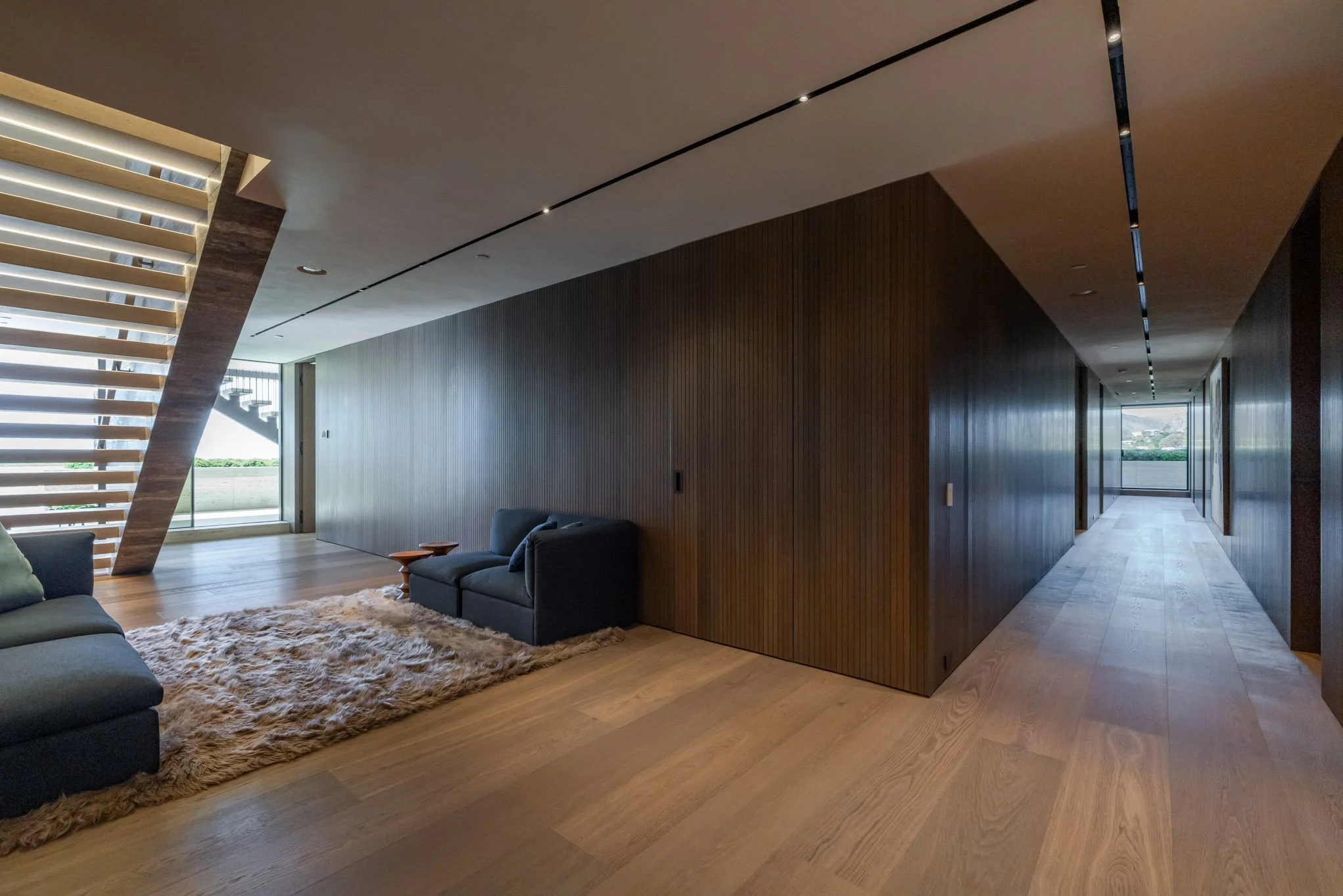 Modern interior living space with wooden flooring, dark sofas, a shaggy rug, a wooden staircase, and large windows at the end of the hallway.