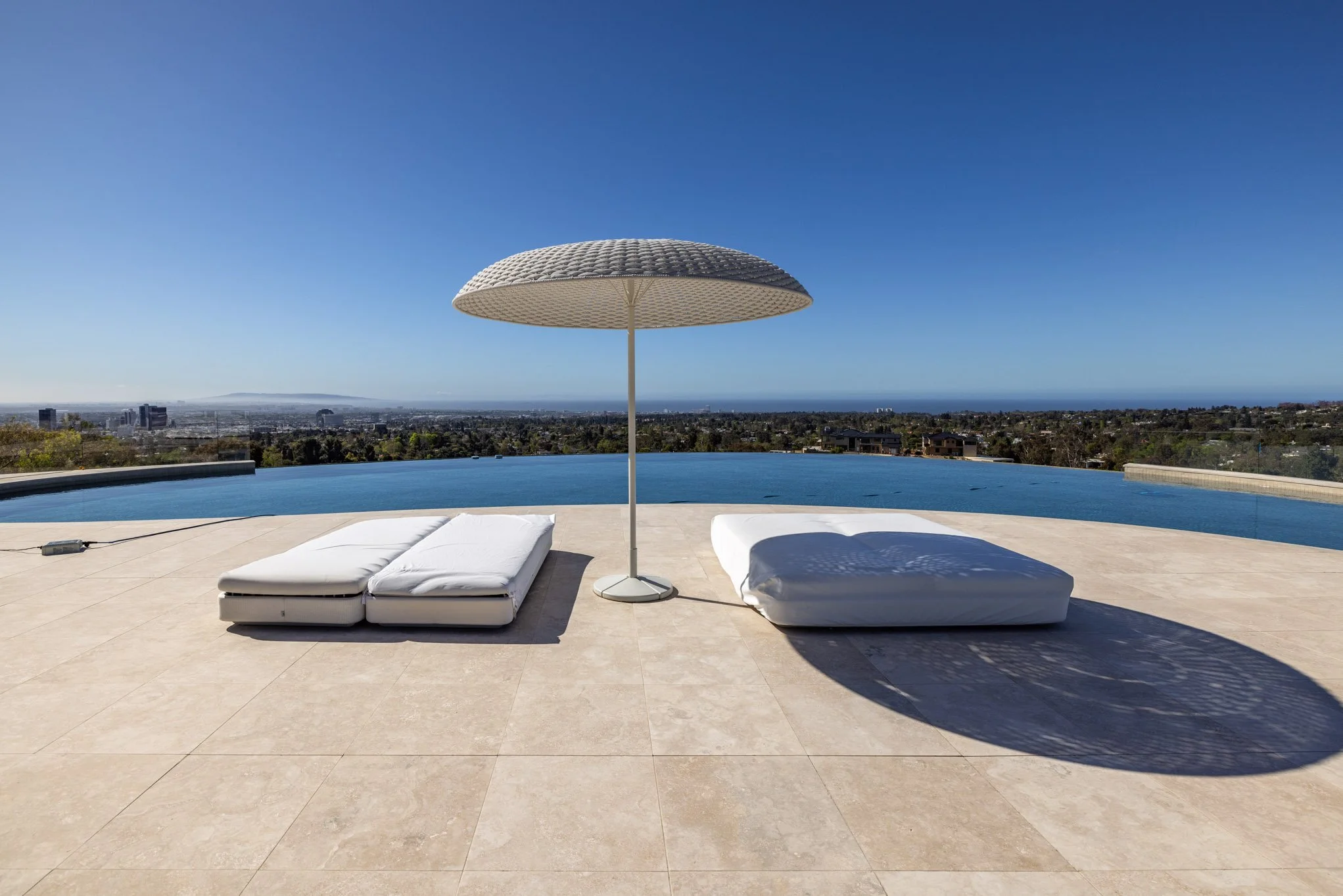 Outdoor pool area with two lounge chairs and a patio umbrella, overlooking a city skyline and ocean view under clear blue sky.