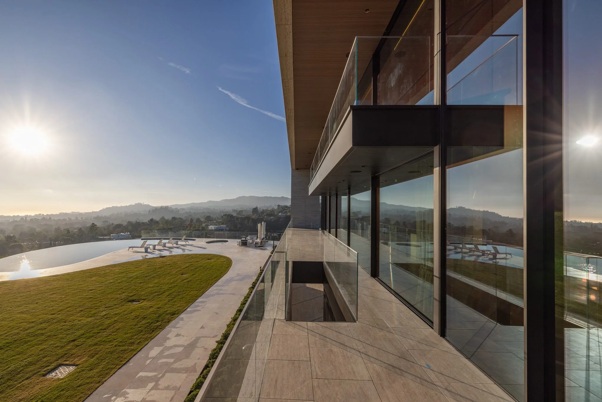 Modern building with large glass windows and a balcony overlooking a landscaped yard with an infinity pool and lounge chairs, hills in the distance, under a clear sky with the sun low on the horizon.