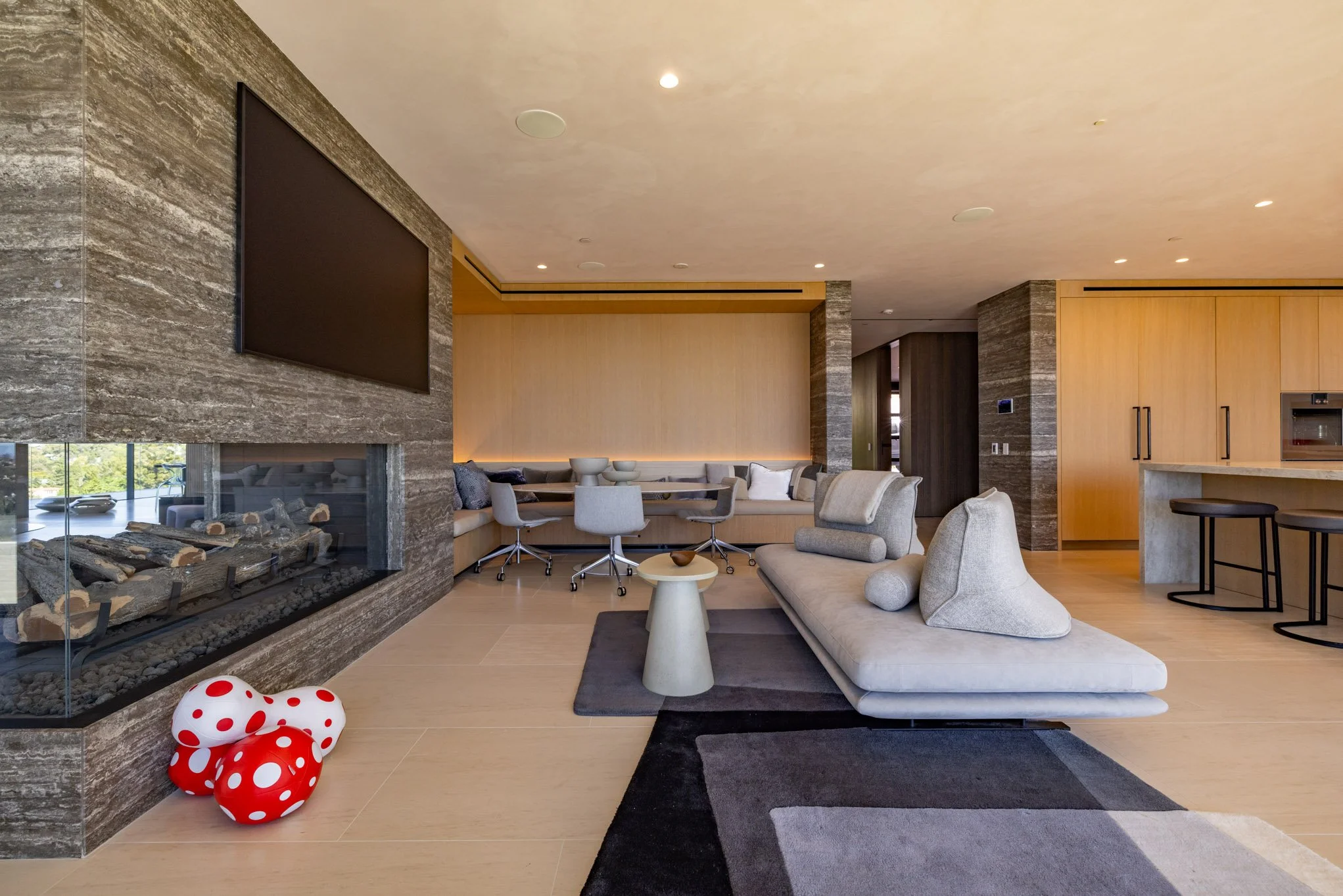 Modern living room with fireplace, light-colored furniture, a flat television, and a kitchenette area with bar stools.
