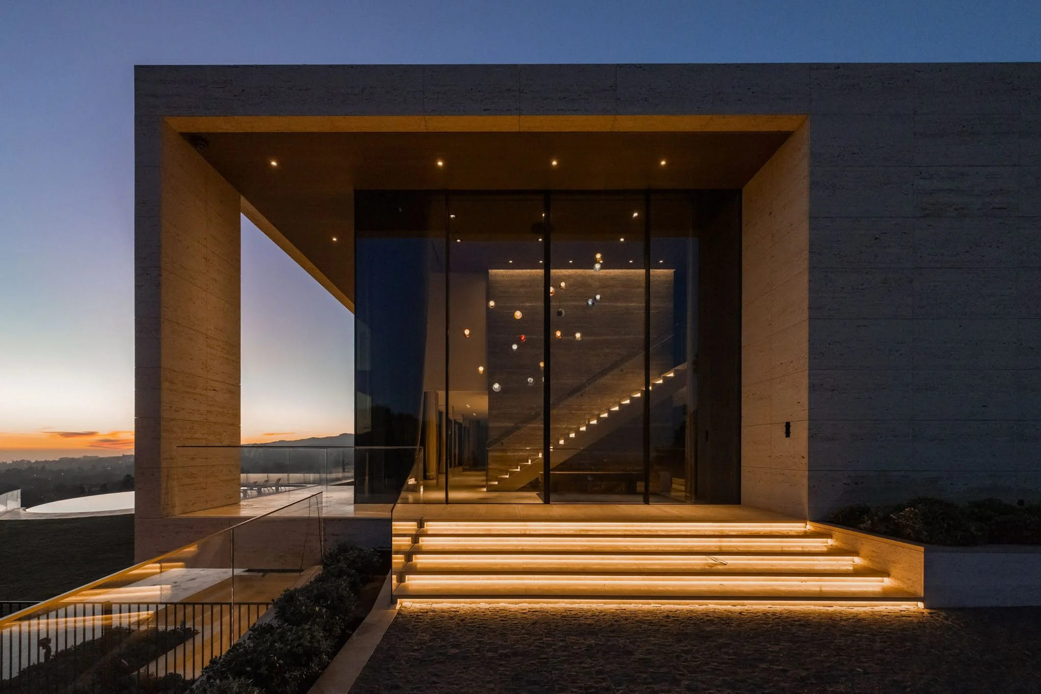 Modern house with a large glass facade, illuminated stairs with warm lighting, during dusk or early evening.