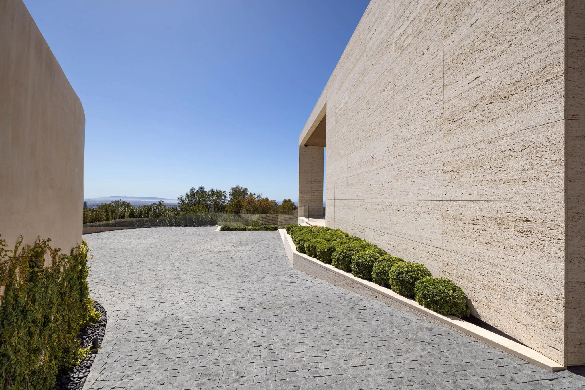 Modern beige stone building with a curved wall, a large rectangular opening, and a row of green bushes along the base, on a cobblestone surface with trees and a clear blue sky in the background.