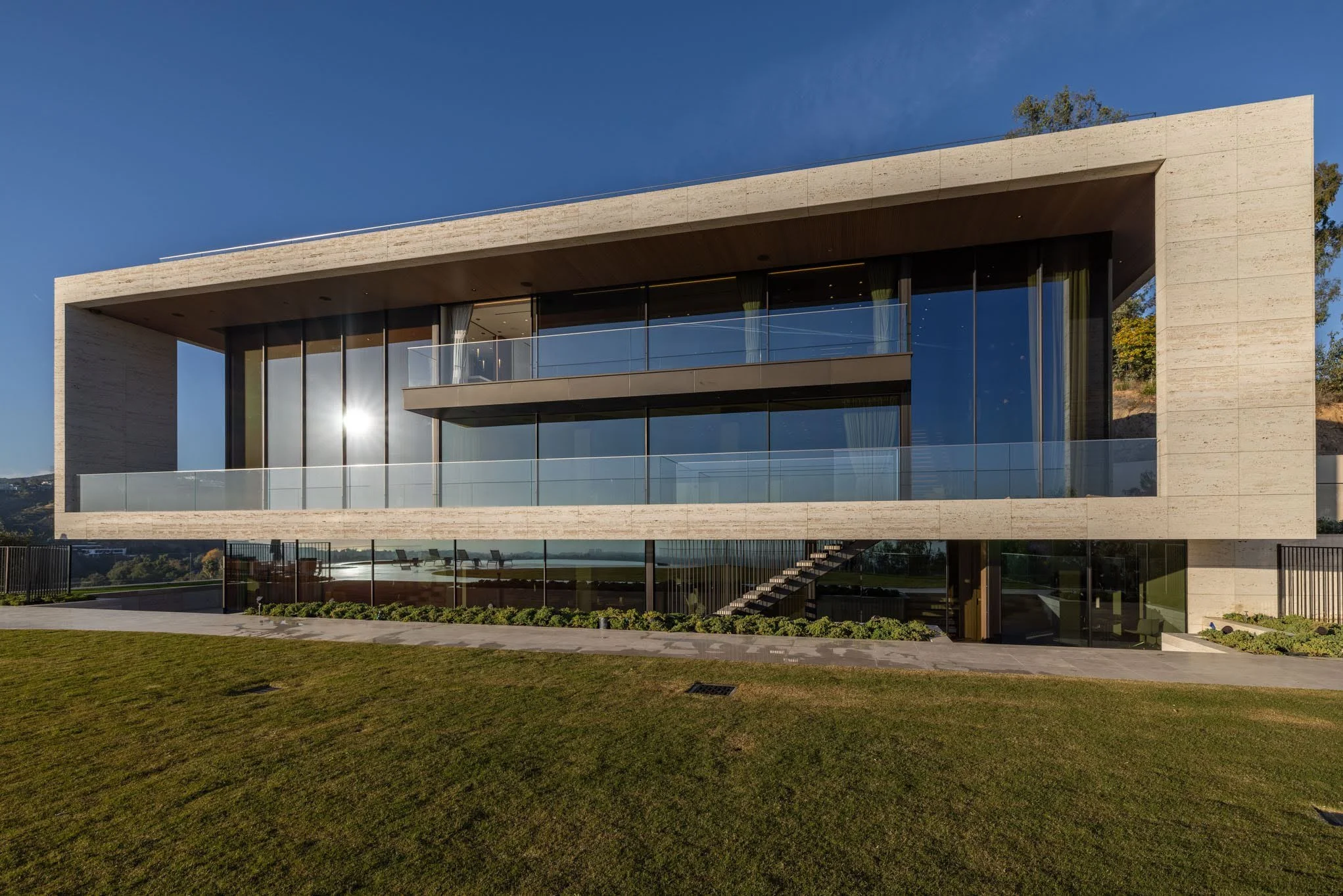Modern two-story house with large glass windows and balconies, surrounded by a well-maintained lawn, with a clear blue sky overhead.