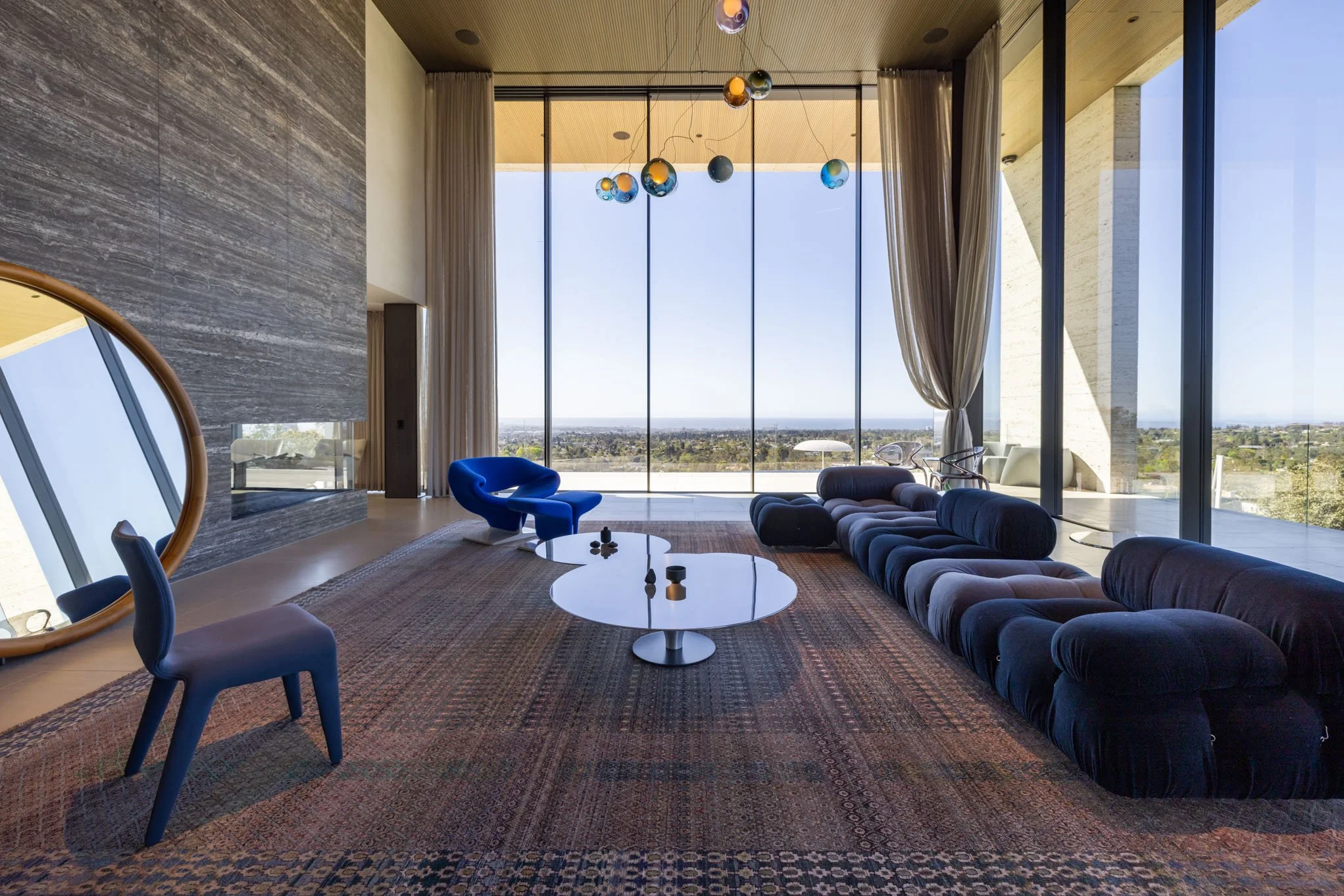 Modern living room with large floor-to-ceiling windows revealing a city view, featuring a long black sectional sofa, a blue accent chair, two white oval coffee tables, and a patterned area rug.
