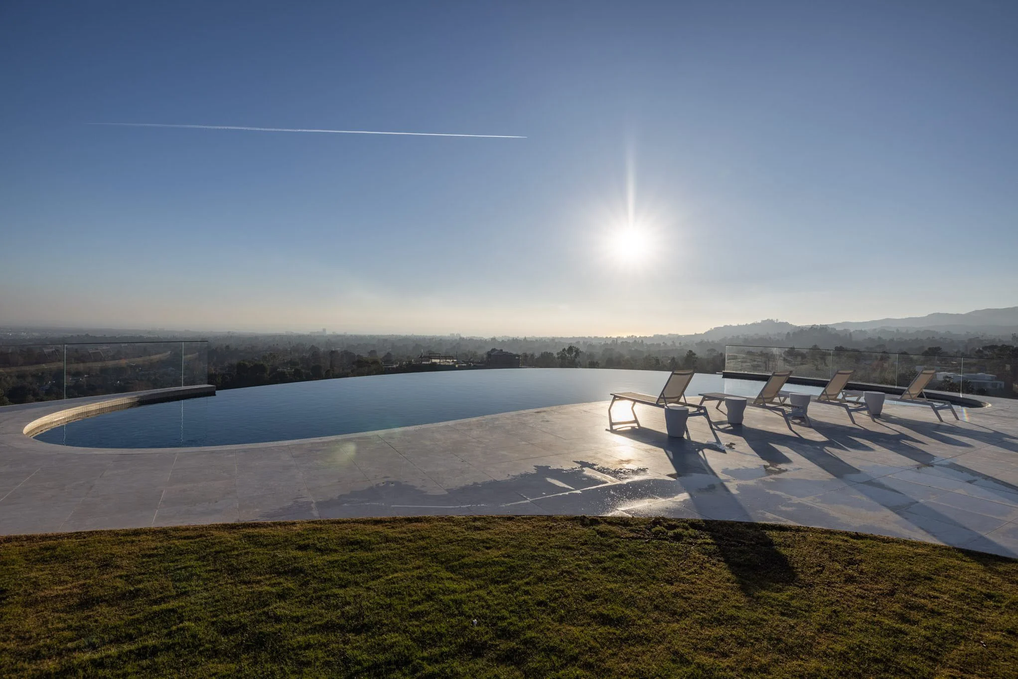 Infinity pool with lounge chairs and small tables on a patio overlooking a cityscape at sunset.