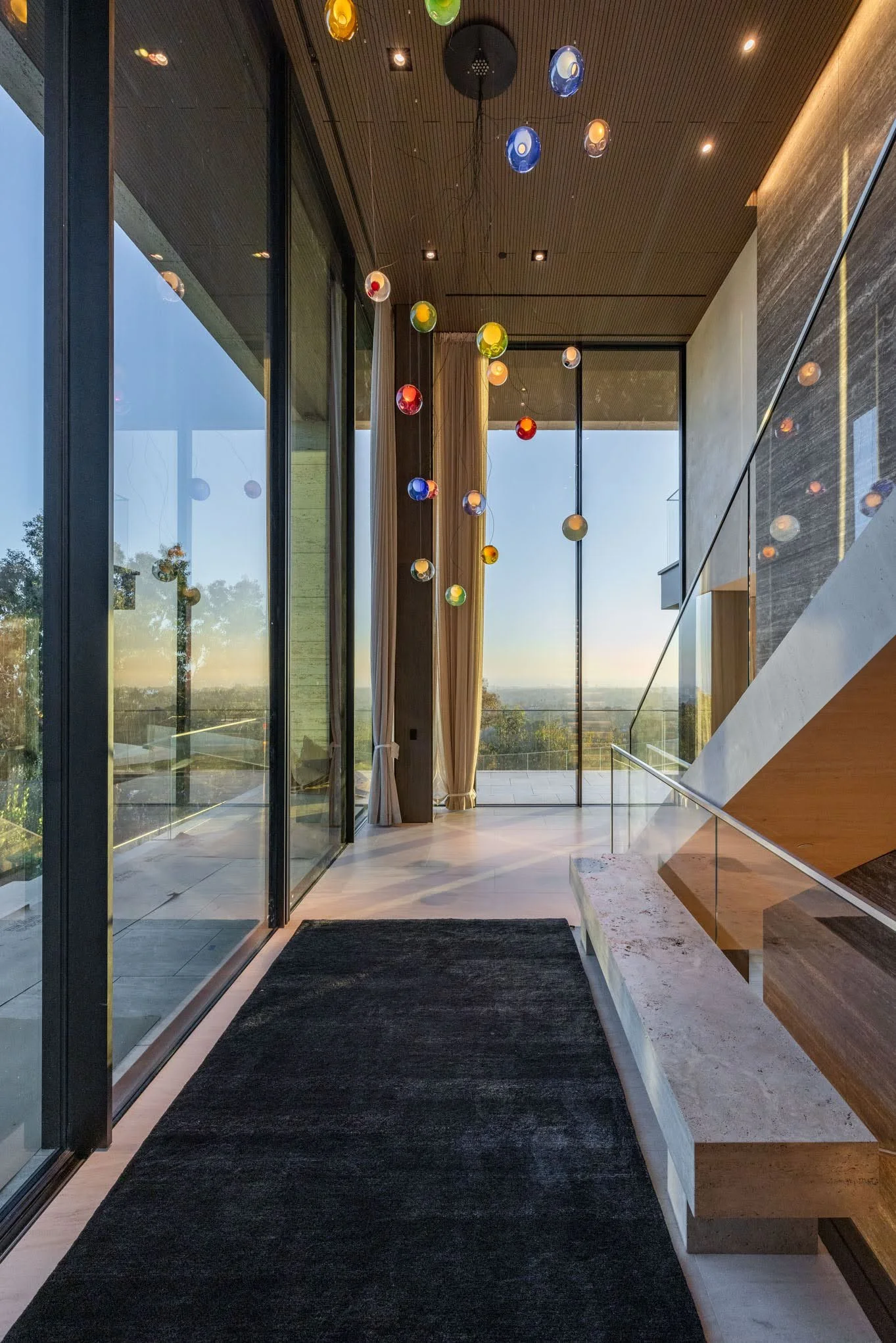 Modern hallway with large glass windows, colorful pendant lights hanging from the ceiling, a black rug, and a view of trees and sky outside.