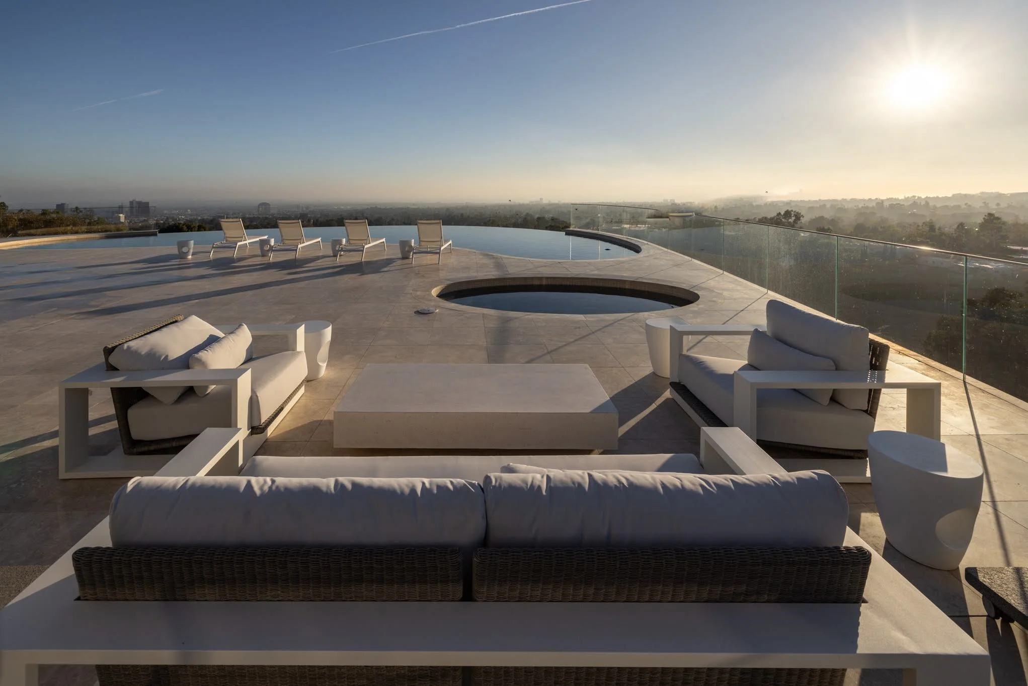 Luxury rooftop terrace with lounge chairs, sofas, and fire pits overlooking a city skyline at sunset.