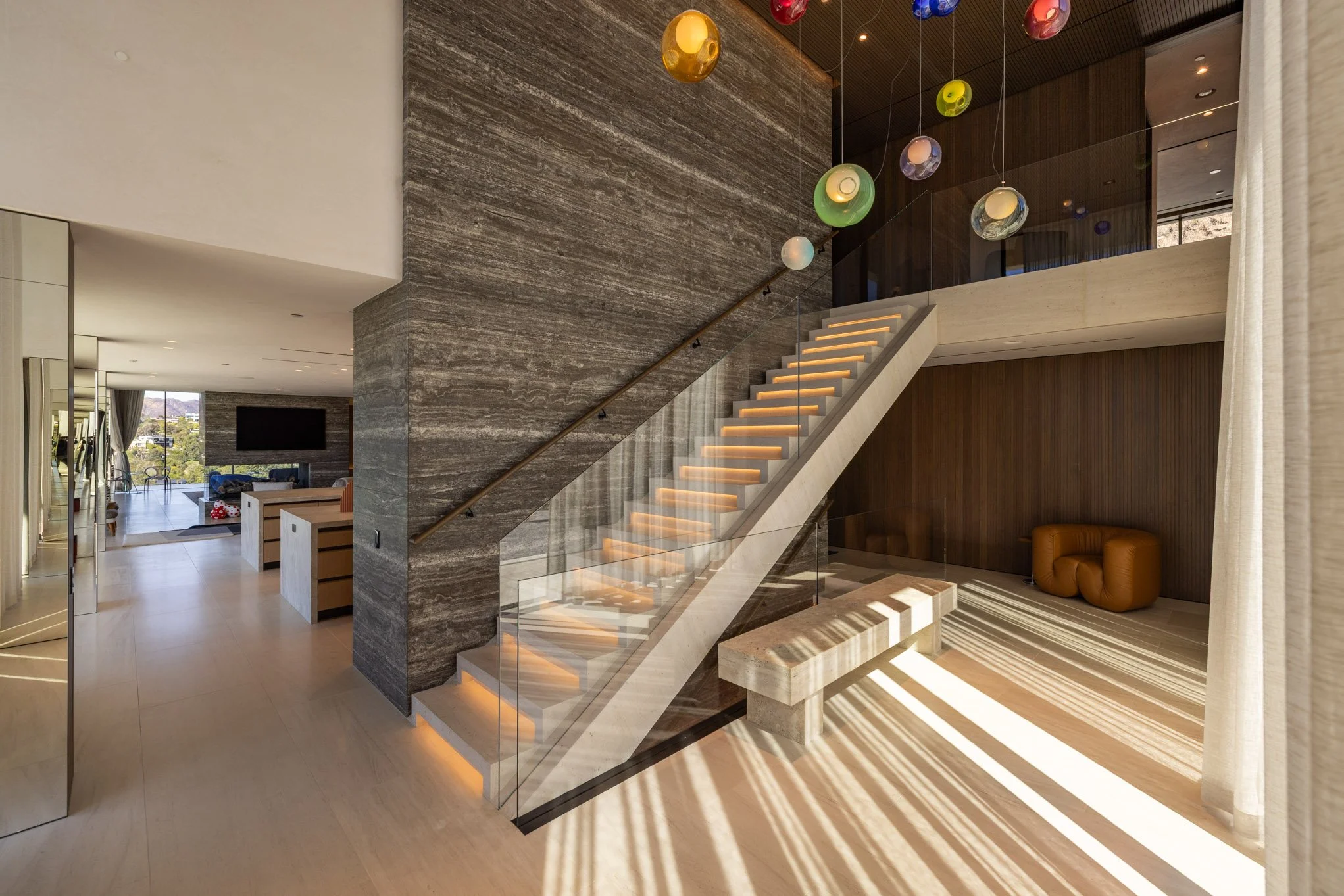 Modern interior of a house featuring a staircase with backlit steps, a seating area with a tan leather ottoman, colorful pendant lights hanging from the ceiling, and a large window letting in natural light.