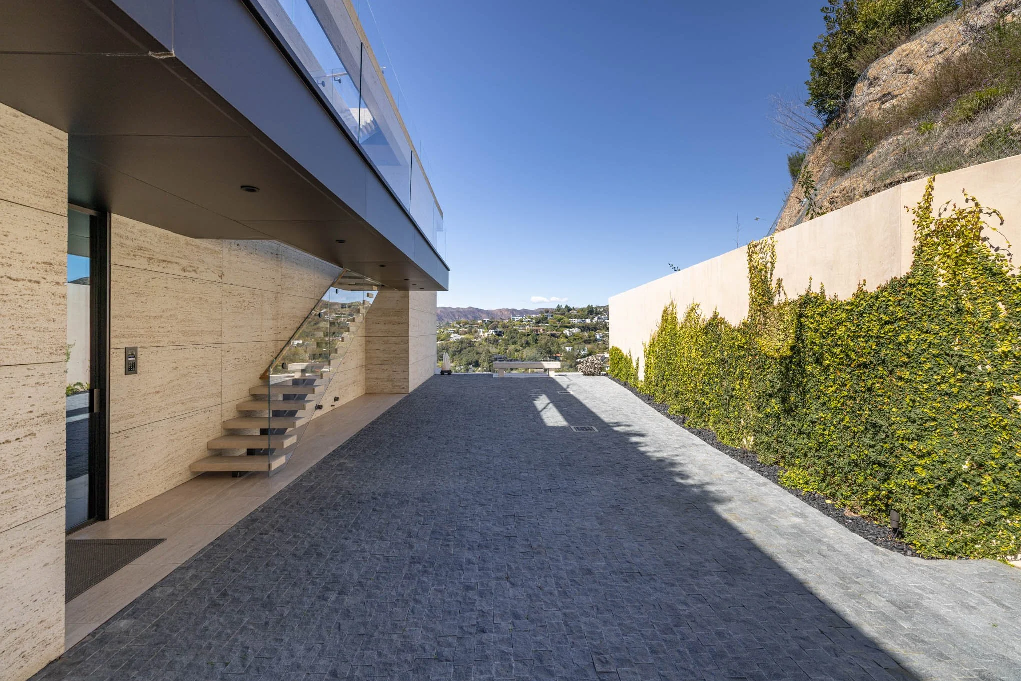 Modern outdoor patio with beige stone wall, glass staircase, and a view of rolling hills in the background. Sunlight casts shadows on the gray cobblestone ground, and a tall green vine-covered wall is on the right.