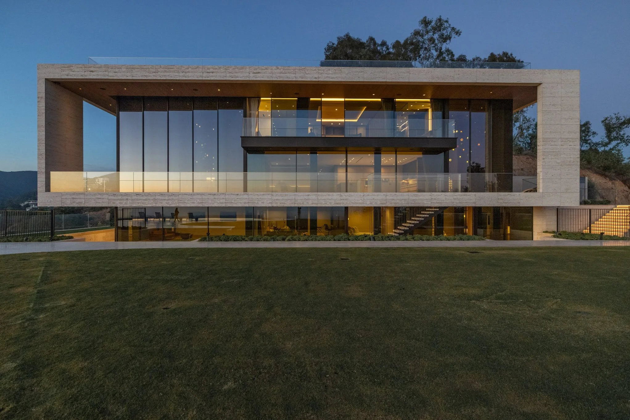 Modern two-story house with large glass windows, illuminated interior, surrounded by a green lawn and trees in the background, during evening