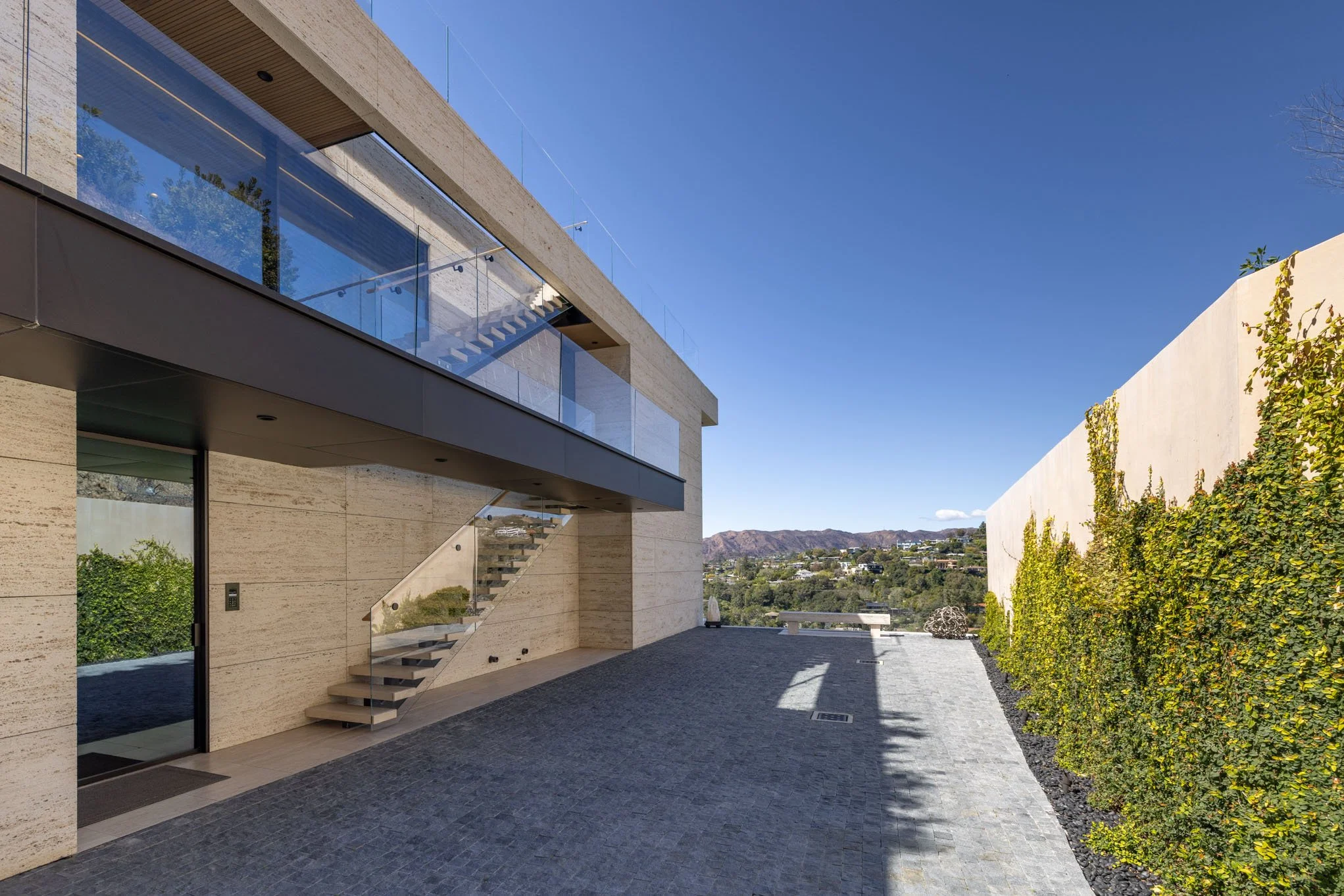 Modern house with beige stone facade, glass balcony, outdoor stairs, gray stone patio, and a tall white wall with green ivy, overlooking a scenic landscape of rolling hills and blue sky.