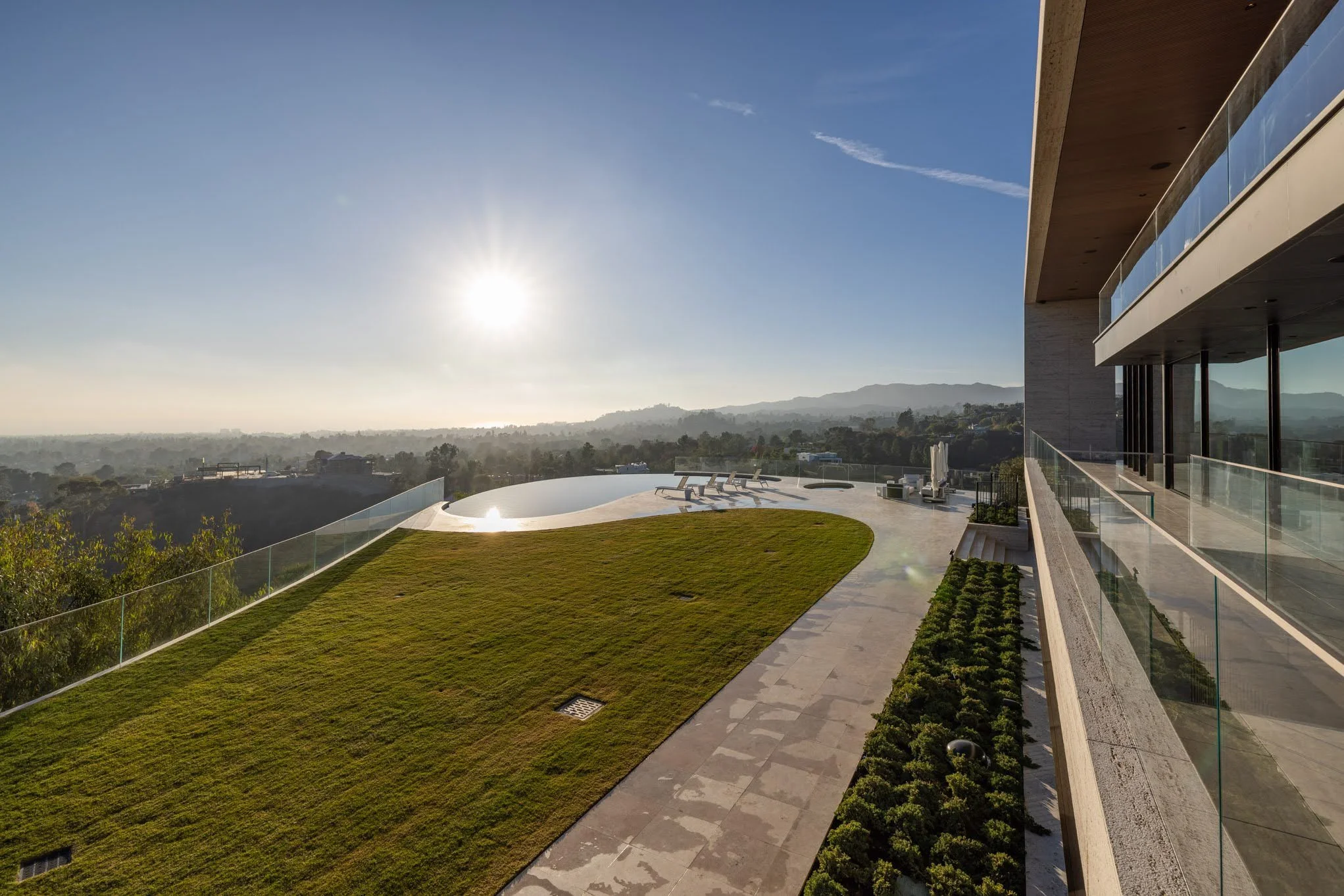 Modern residential balcony with green lawn, glass railing, sun shining, overlooking a scenic landscape with hills in the distance.